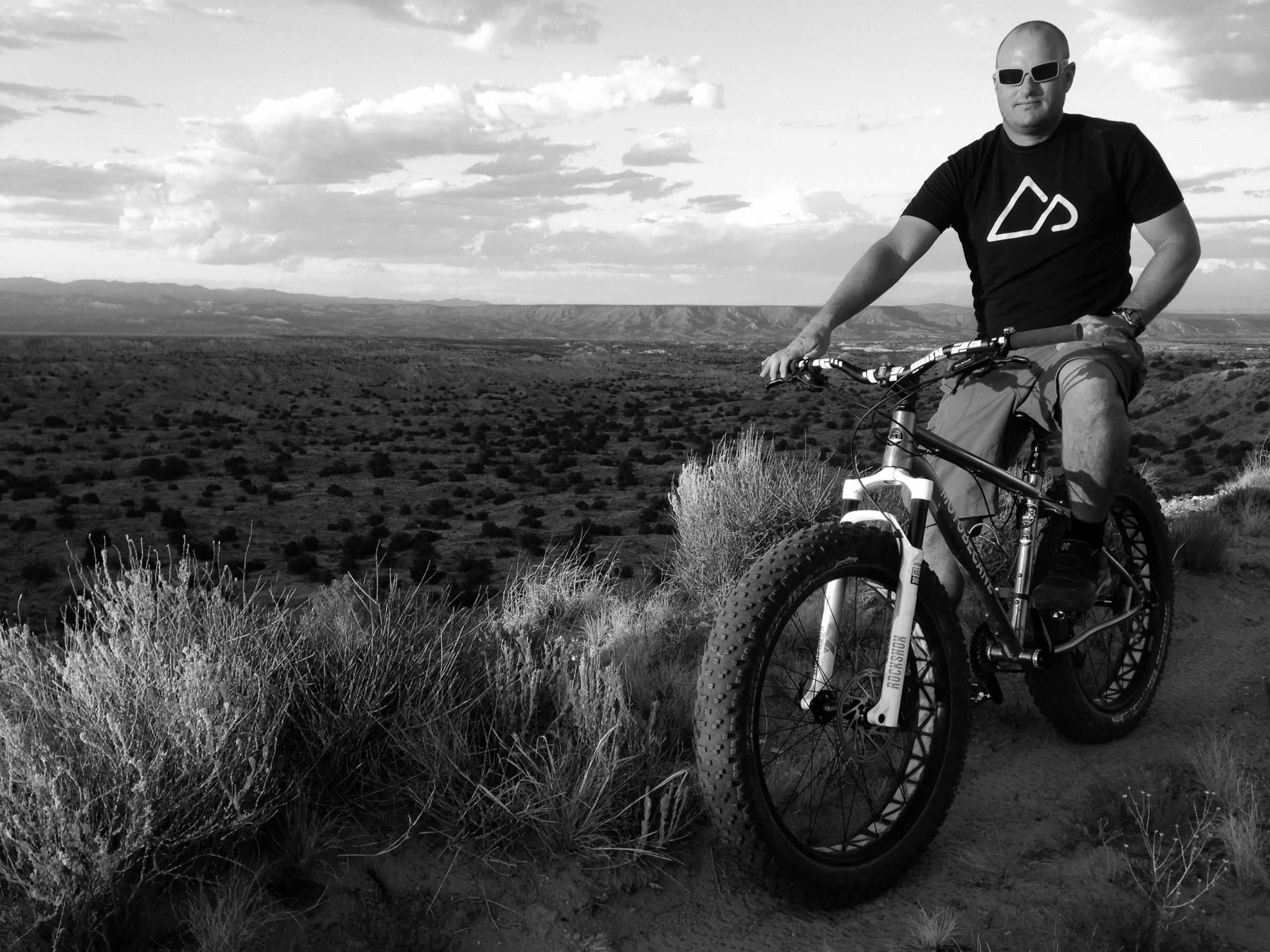 A cyclist wearing sunglasses and a black t-shirt sits on a mountain bike on a rocky trail overlooking a vast, mountainous landscape. The image is in black and white, showcasing a rugged terrain with sparse vegetation and a cloudy sky. Mariposa Fat Bike Trails mountain bike trail.