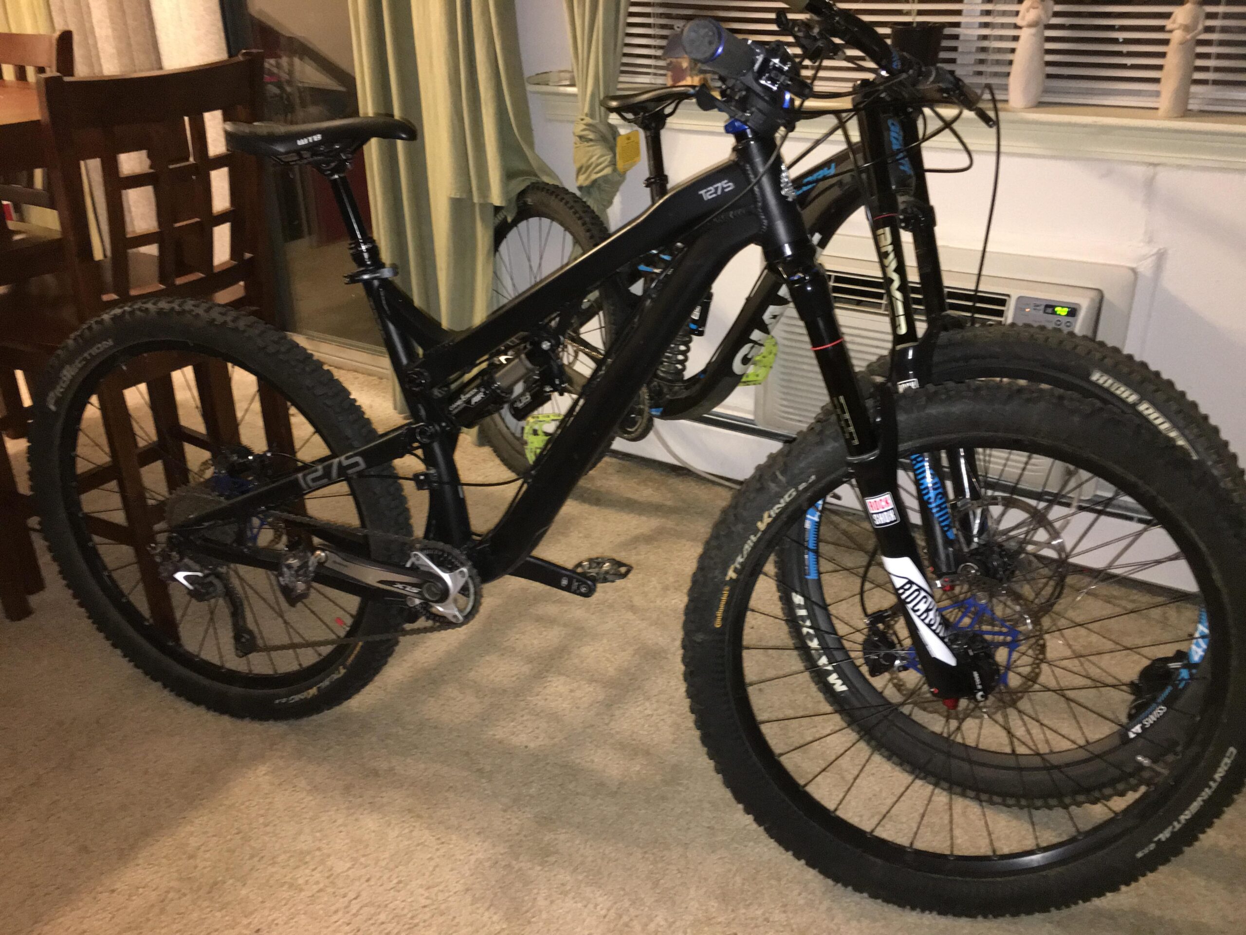 Intense tracer: A black mountain bike with thick tires is leaning against a chair in a living room setting. The bike features a dropper seatpost and visible gears, with another bike partially in the background. The room has light-colored walls, curtains, and a small air conditioning unit on the side.