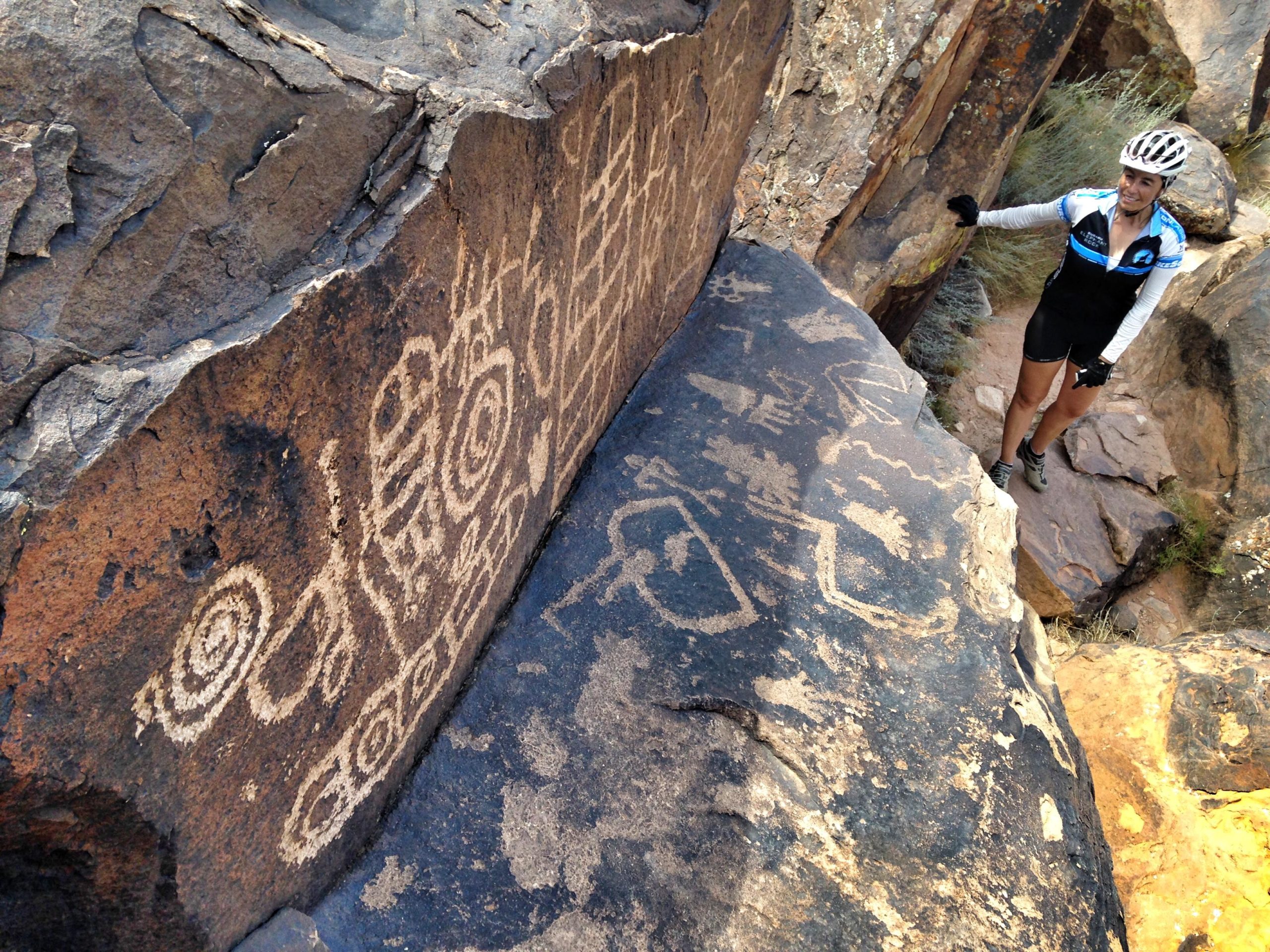 A person in cycling attire stands next to large rock formations displaying ancient petroglyphs, which include spiral and geometric designs. The scene is set in a rugged outdoor environment with rocky surfaces and sparse vegetation. Barrel Roll mountain bike trail.