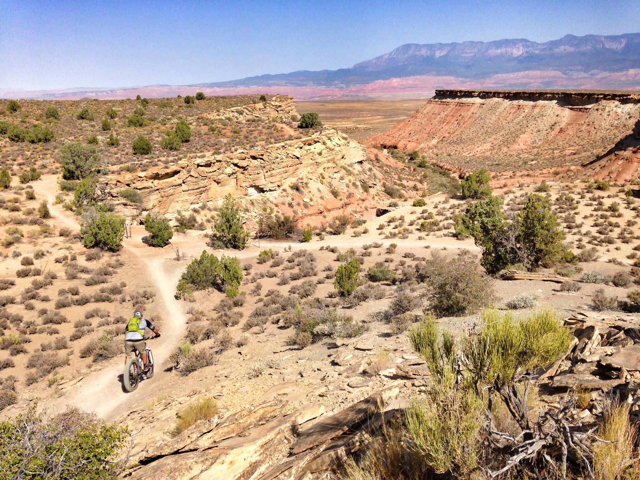 A mountain biker rides down a dirt trail through a rugged desert landscape, surrounded by sparse vegetation and rocky cliffs under a clear blue sky, with distant mountains visible on the horizon. J.E.M. Trail mountain bike trail.