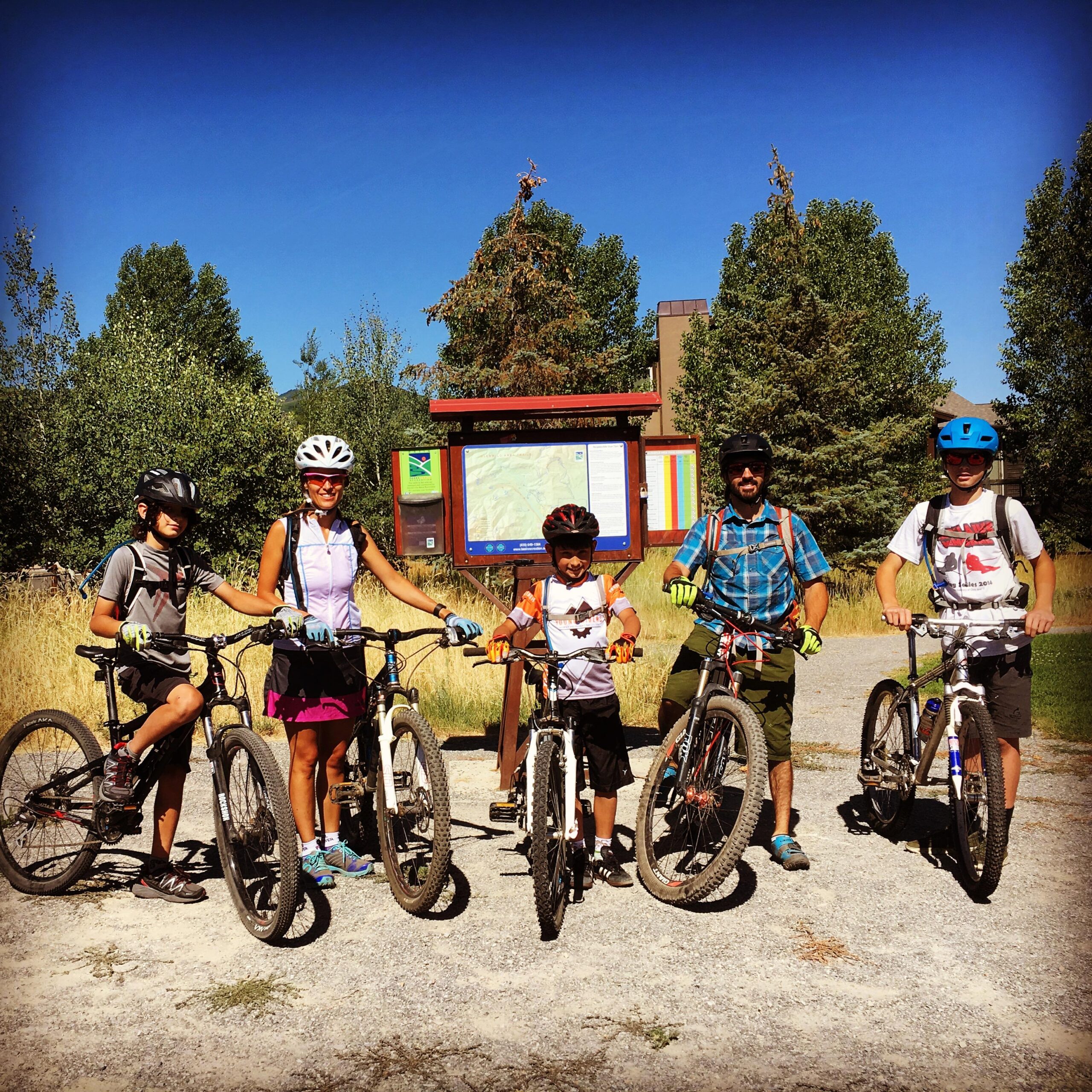 Giant Anthem X 29er 1: A family of five stands with mountain bikes in front of a trail map sign in a grassy outdoor area. The group includes two boys and a girl, all wearing helmets and cycling gear, with bright sunlight and clear blue skies in the background. Trees line the background, suggesting a recreational area for biking.