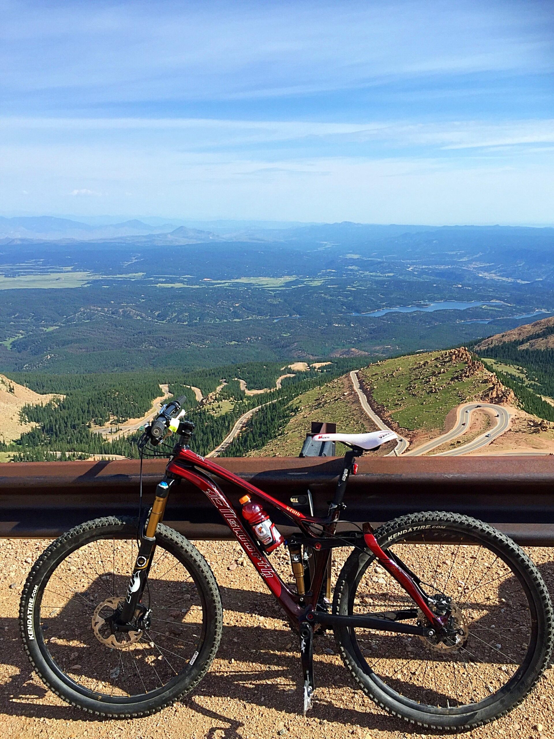 Ellsworth Truth: A mountain bike parked at a scenic viewpoint with a panoramic view of rolling hills and a winding road below, under a clear blue sky. The bike is red with black accents and has a water bottle mounted on the frame.