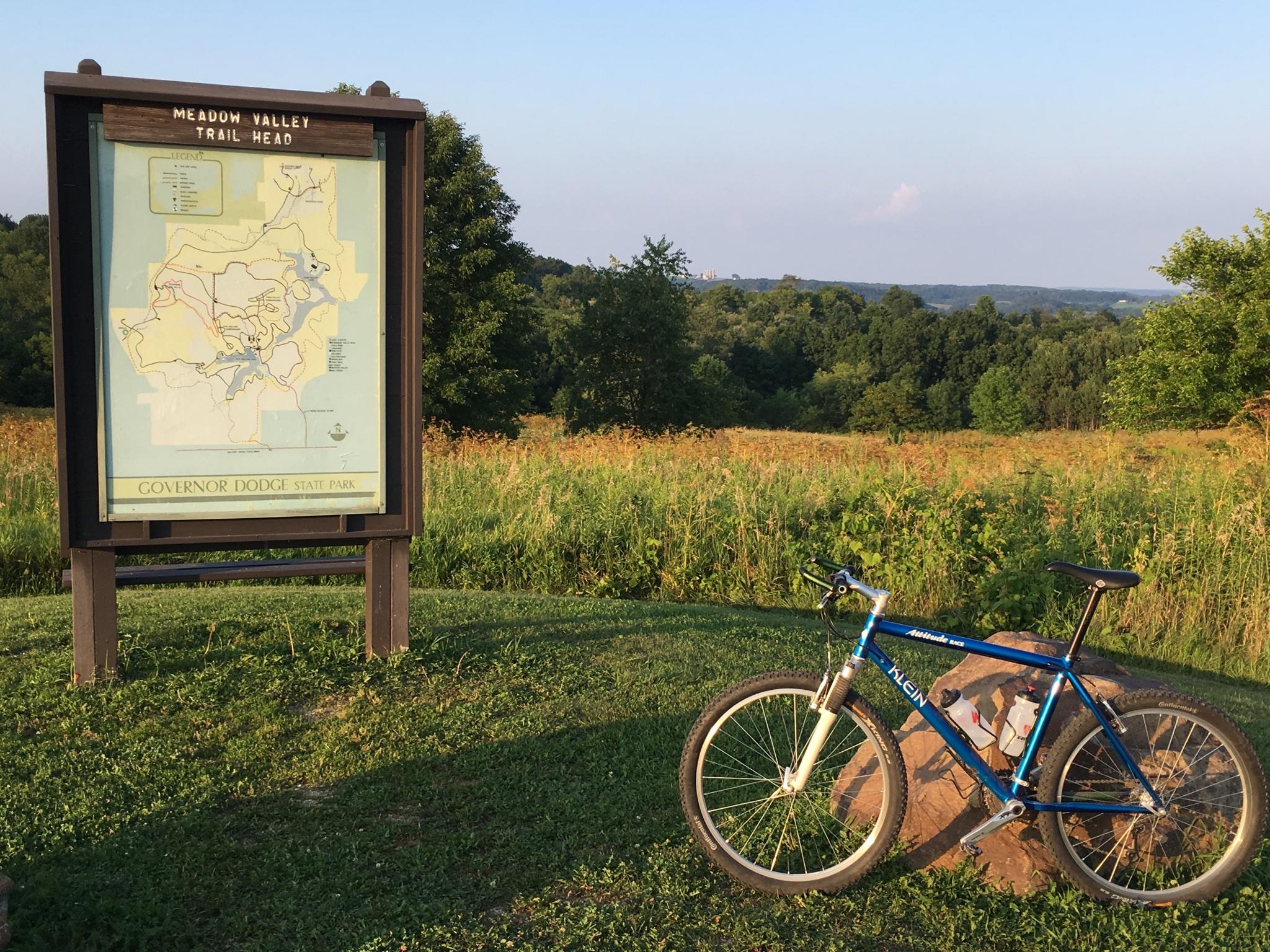 Klein Attitude Race: Trail head sign and bike at Governor Dodge State Park. The sign, titled "Meadow Valley Trail Head," displays a map of the park's trails. A blue mountain bike rests on a rock in the foreground, with a backdrop of lush green fields and distant hills under a clear sky.