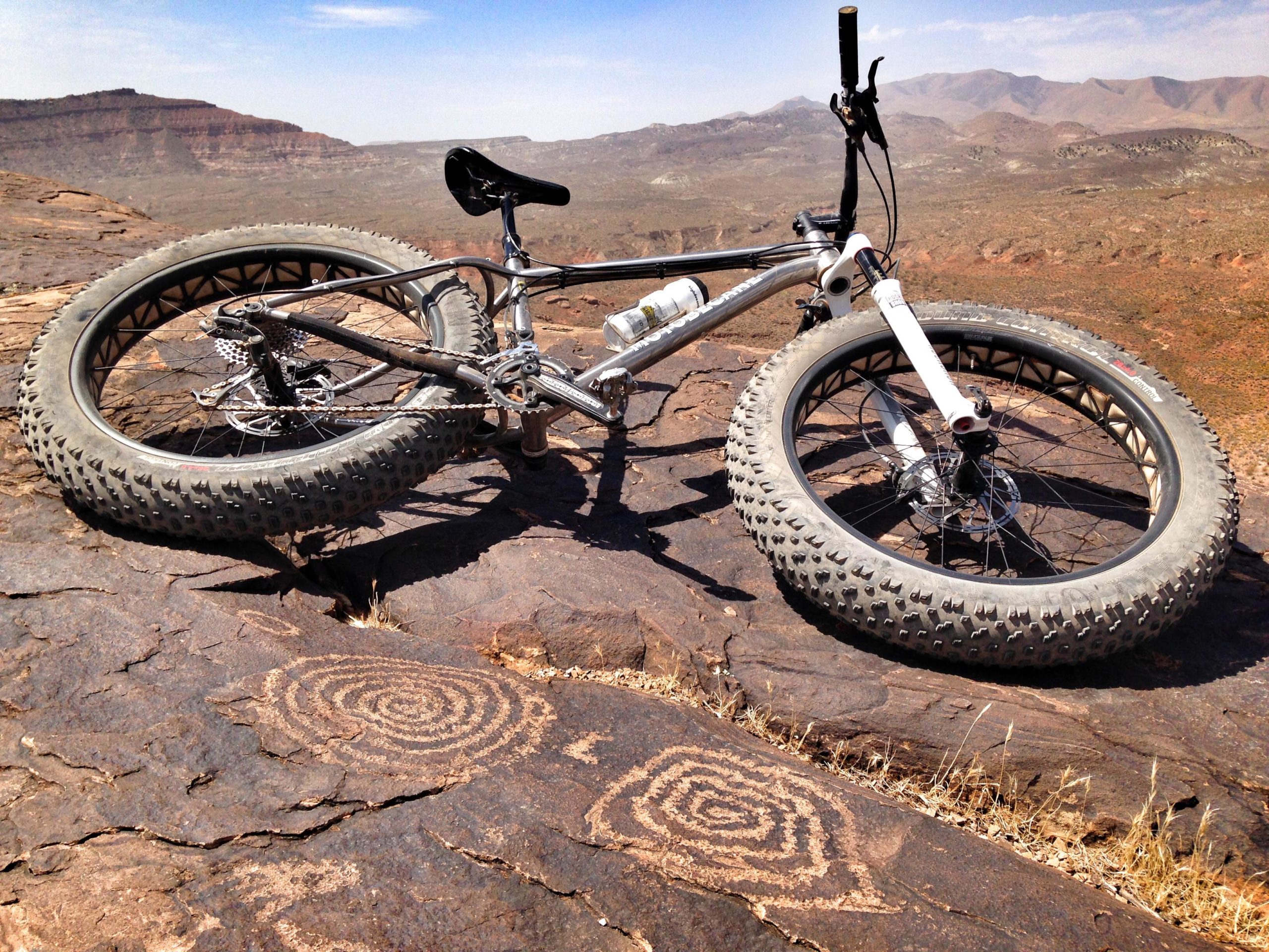 A fat tire bicycle resting on rocky terrain, with petroglyphs featuring spiral designs etched into the stone. In the background, a scenic view of distant mountains and a clear blue sky. Barrel Roll mountain bike trail.