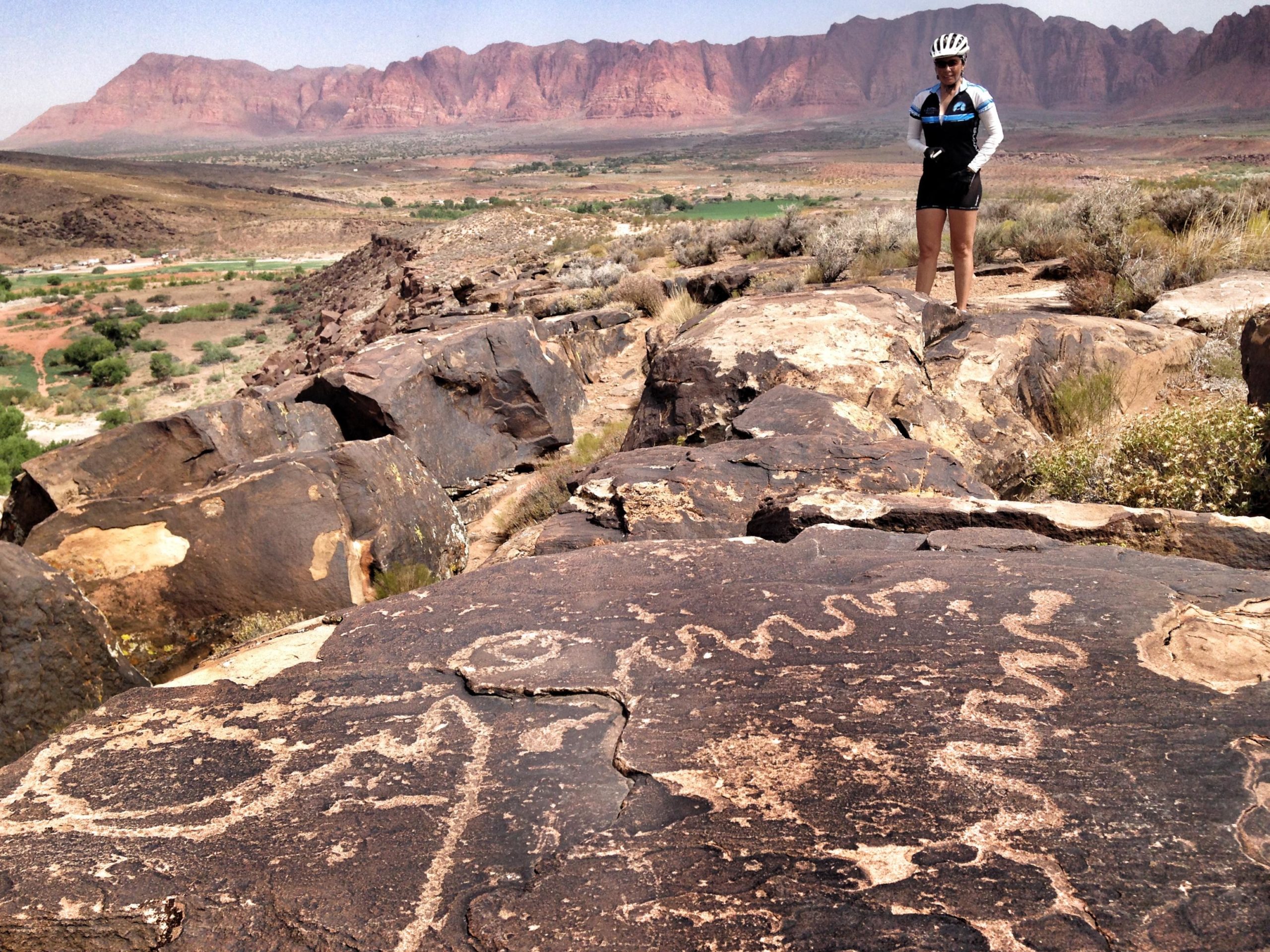 A woman in a cycling outfit stands on a rocky outcrop, observing ancient petroglyphs carved into the stones. The surrounding landscape features red mountains in the background, with patches of green vegetation in the valley below. The scene captures a blend of natural beauty and historical artistry. Barrel Roll mountain bike trail.