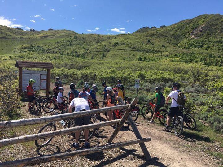 Giant Anthem X 29er 1: A group of mountain bikers gathered at the trailhead, surrounded by lush greenery and rolling hills. They are wearing helmets and colorful biking gear, with some adjusting their bikes. A wooden sign is visible in the background, indicating trail information, and a wooden fence lines the foreground. The sky is bright and clear, suggesting a sunny day.