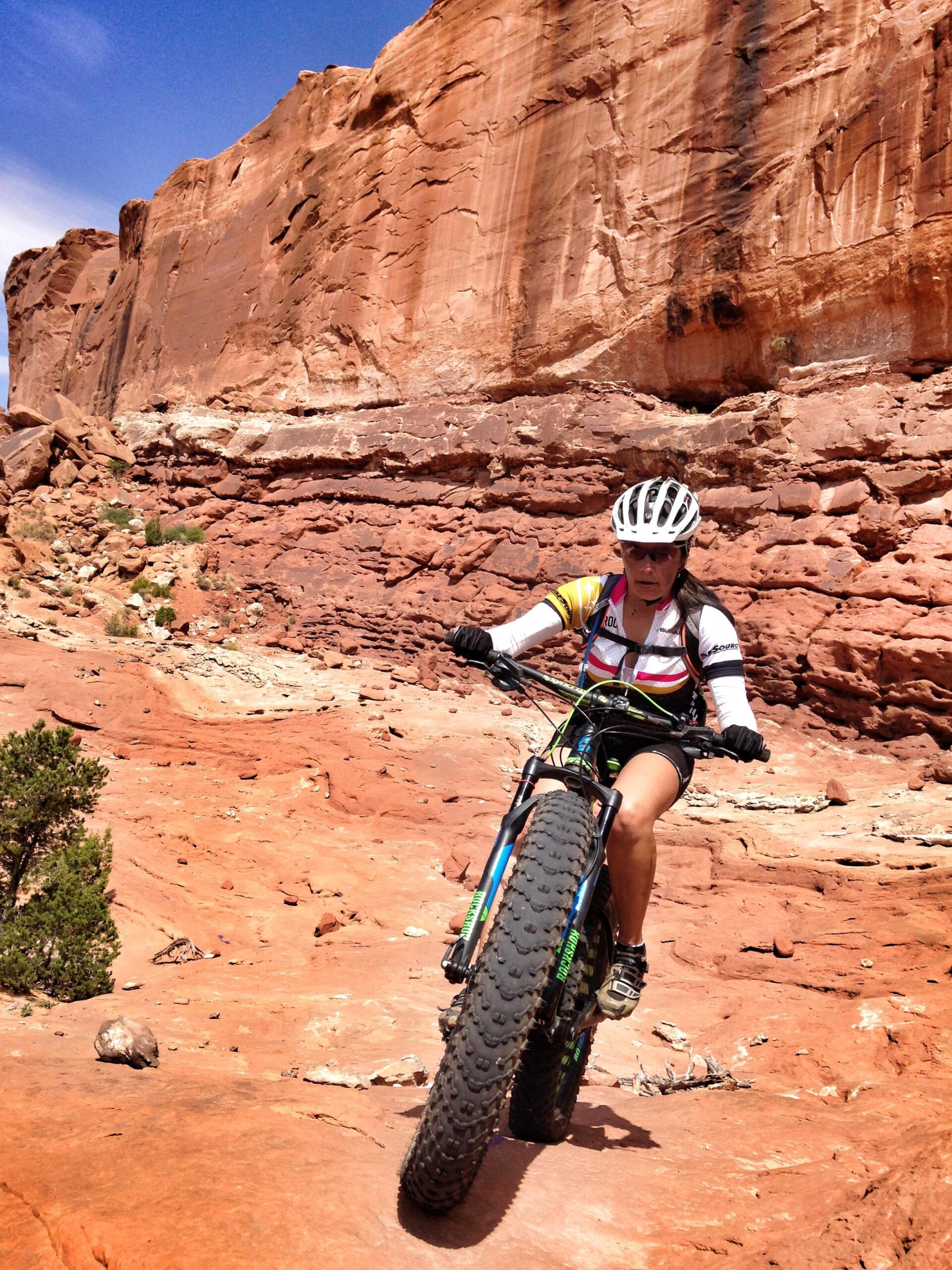 A mountain biker riding over rocky terrain in a desert landscape, with red rock formations in the background and a clear blue sky overhead. The biker is wearing a helmet, gloves, and a colorful jersey, demonstrating an advanced biking maneuver. Navajo Rocks mountain bike trail.