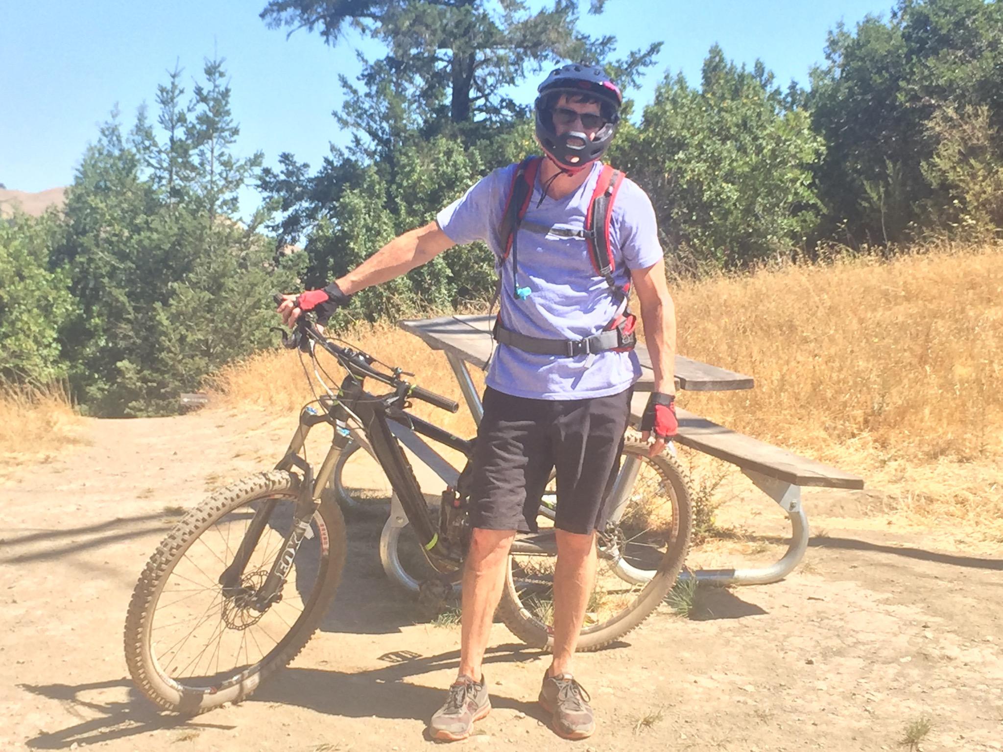 A person wearing a helmet and biking gear stands beside a mountain bike on a dirt path surrounded by trees and dry grass. The individual is wearing a blue shirt, black shorts, and red gloves, and is posed next to a picnic table in a natural outdoor setting. Camp Tamarancho mountain bike trail.