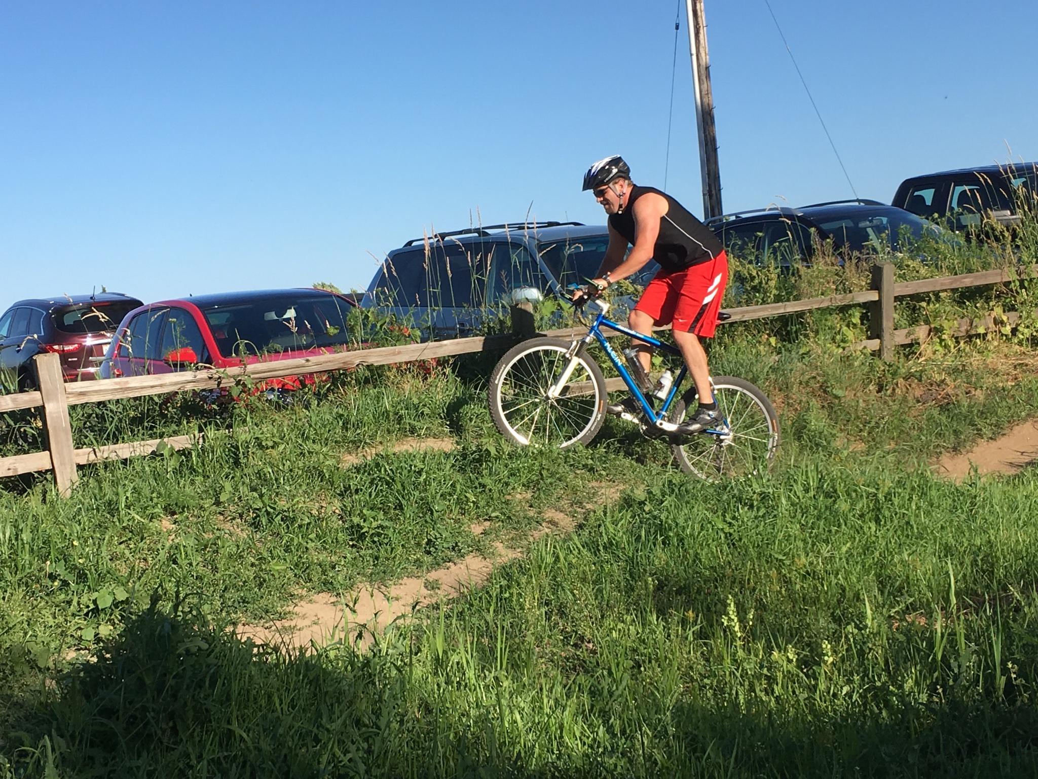 Klein Attitude Race: A man in a sleeveless black shirt and red shorts rides a mountain bike on a grassy trail near a wooden fence, with parked cars visible in the background under a clear blue sky.