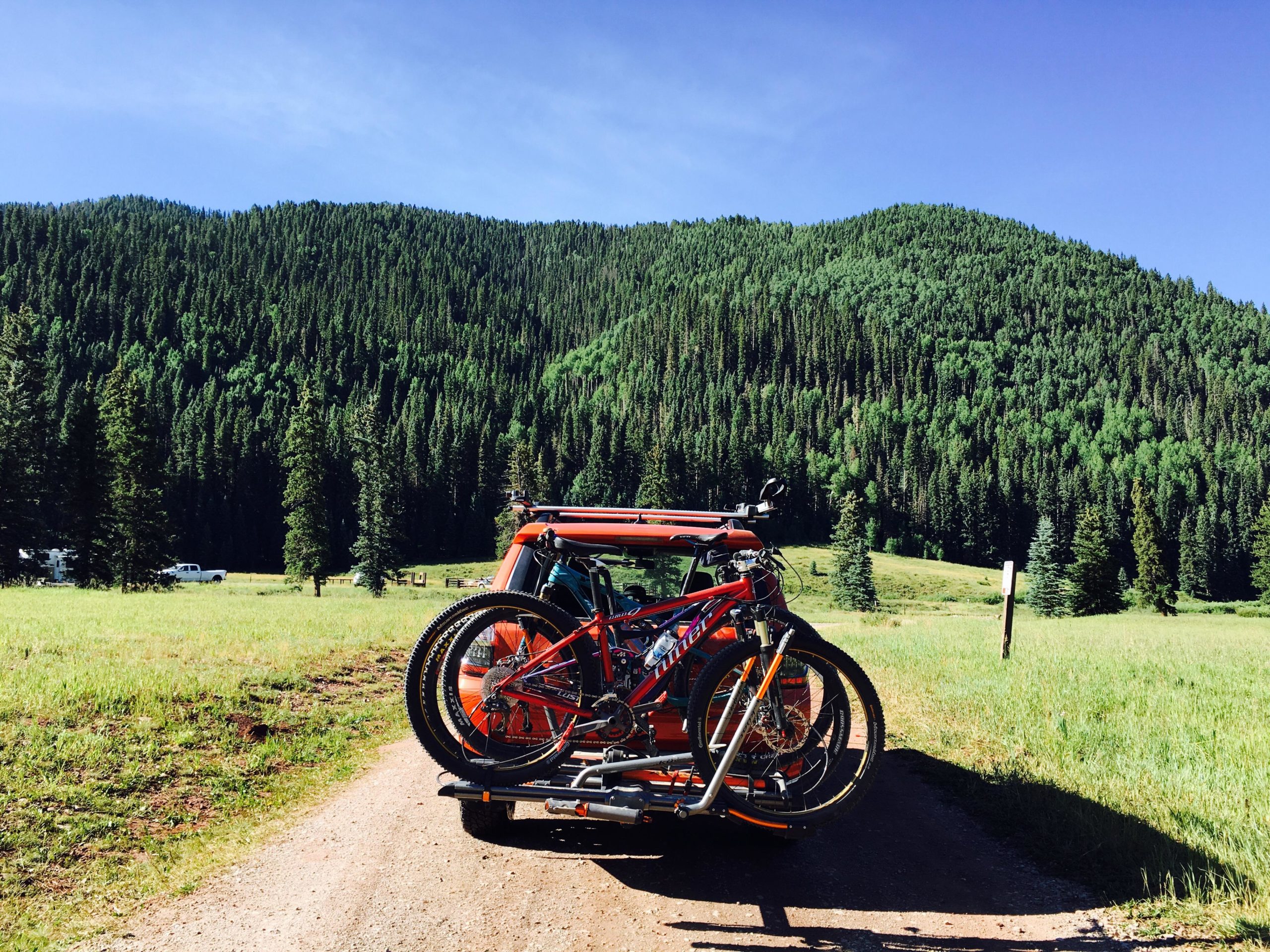 A red vehicle parked on a dirt road with two mountain bikes mounted on a bike rack, surrounded by a lush green meadow and dense pine trees in the background under a clear blue sky. Hermosa Creek Trail mountain bike trail.