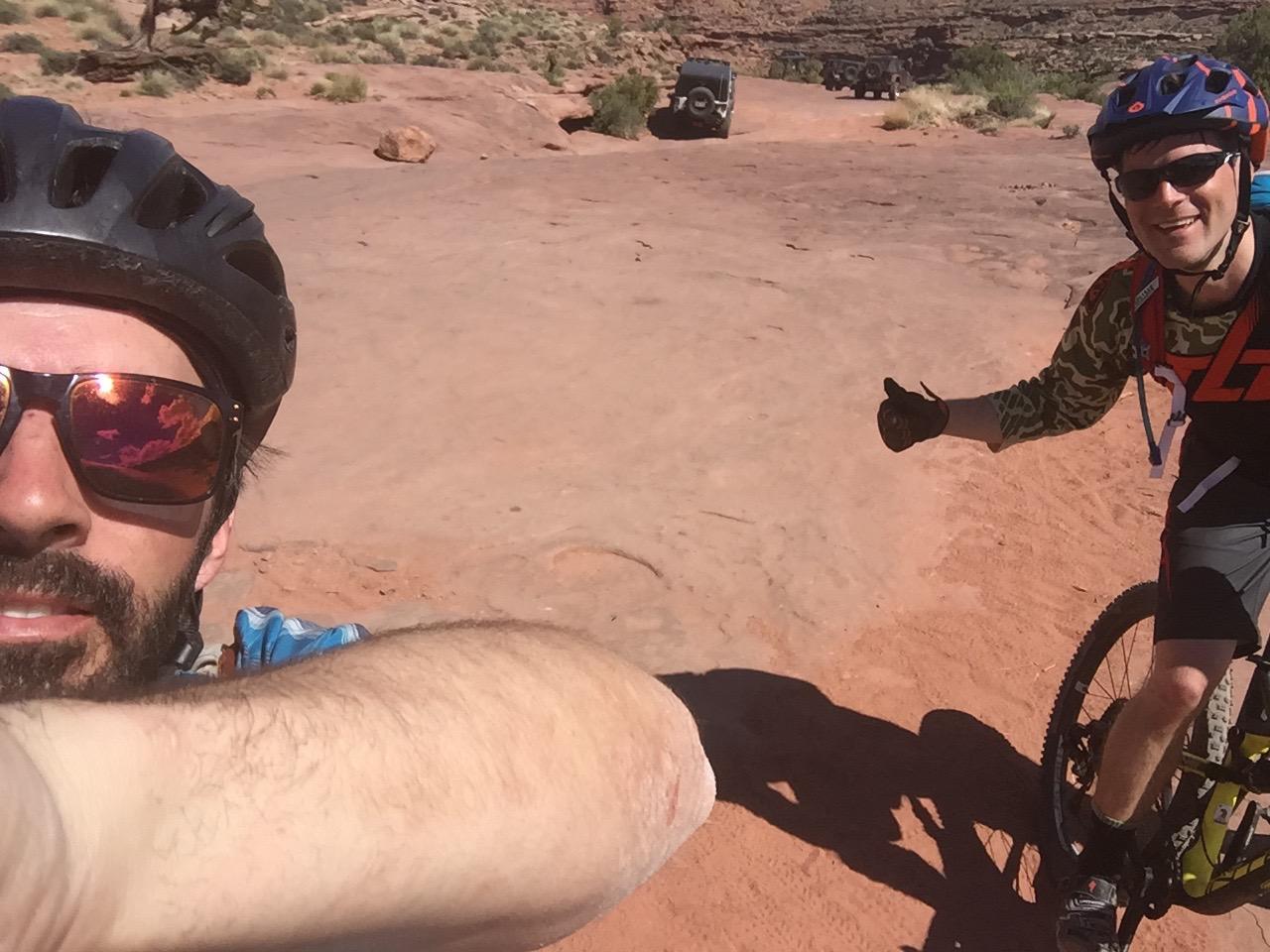 Giant Anthem X 29er 1: Two mountain bikers take a selfie on a rocky terrain under bright sunlight. One biker, wearing a black helmet and sunglasses, is visible prominently in the foreground, while the other, in a camo-patterned shirt, gives a thumbs-up in the background. Off-road vehicles are parked in the distance, surrounded by a natural landscape of red rock and sparse vegetation.