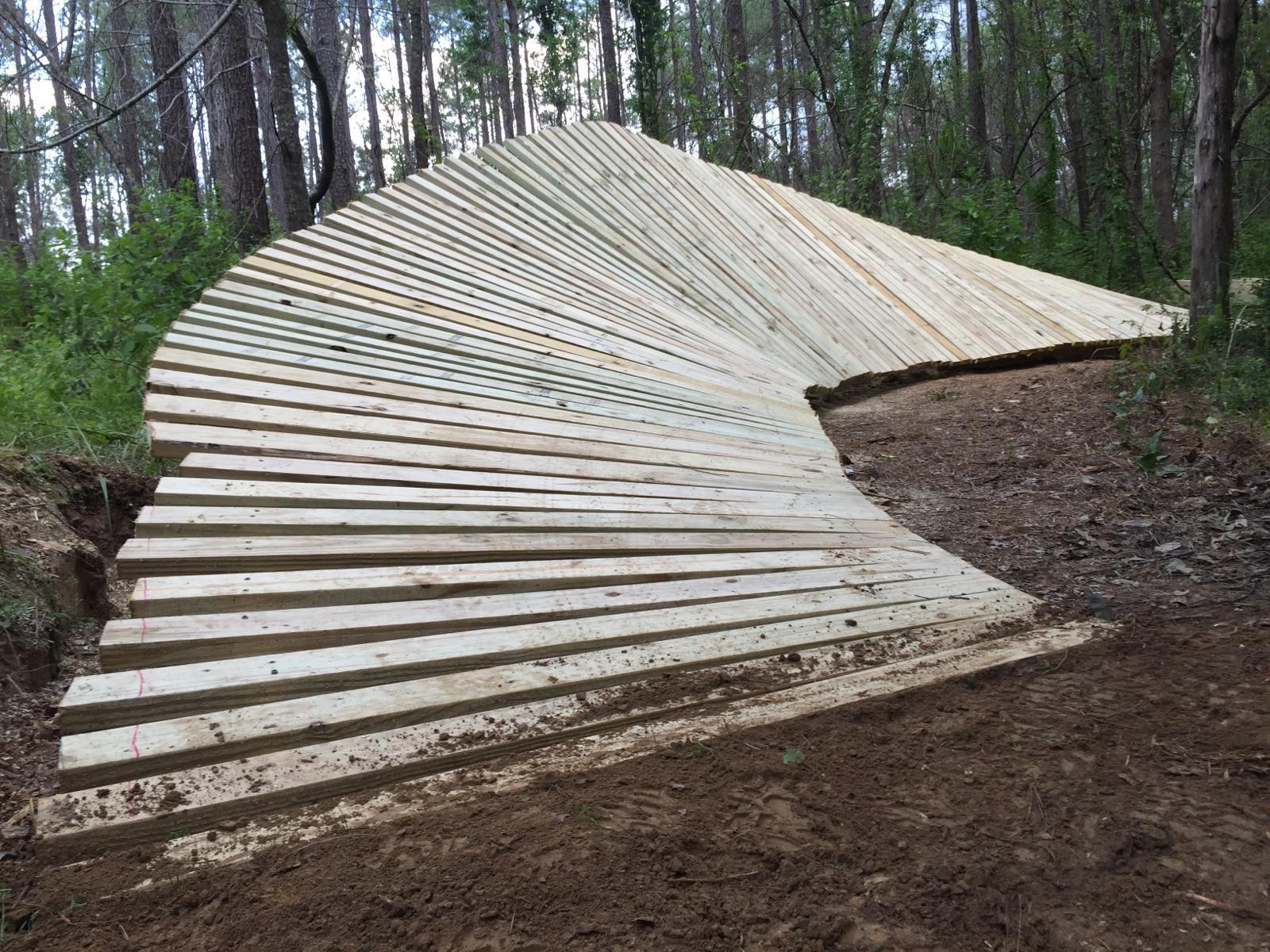 A wooden ramp featuring a curved design, constructed from slats arranged in a spiral pattern. The ramp is situated in a forested area, surrounded by trees and underbrush. The ground beneath the ramp is bare soil, showing signs of recent excavation. Mt. Zion Bike Trails mountain bike trail.