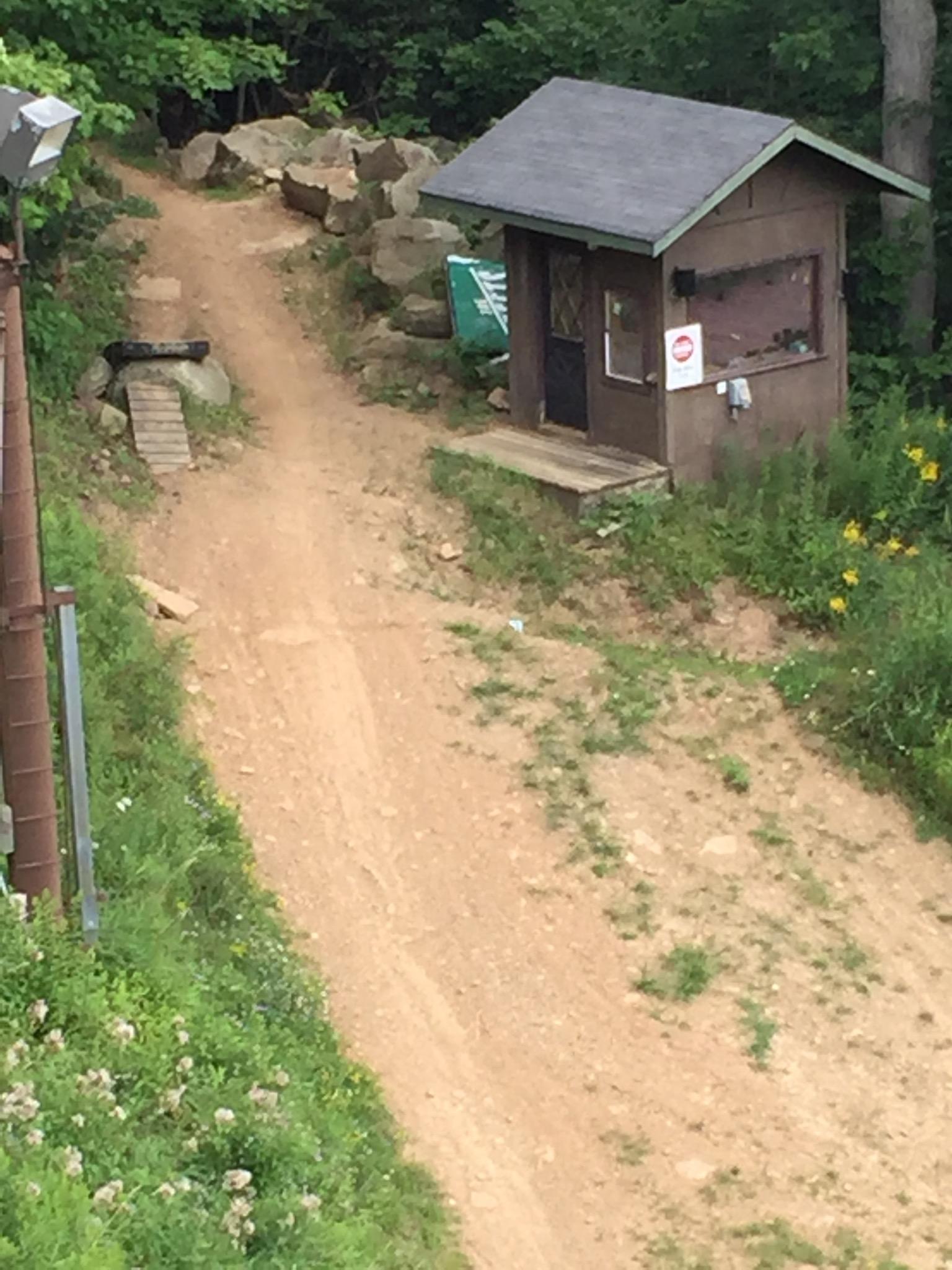 A dirt path leading up a hill, bordered by rocks and greenery. On the right side of the path, there is a small wooden shed with a dark door and a window. The shed appears rustic, with some signage visible. Surrounding the area are patches of wildflowers and lush foliage. Seven Springs mountain bike trail.