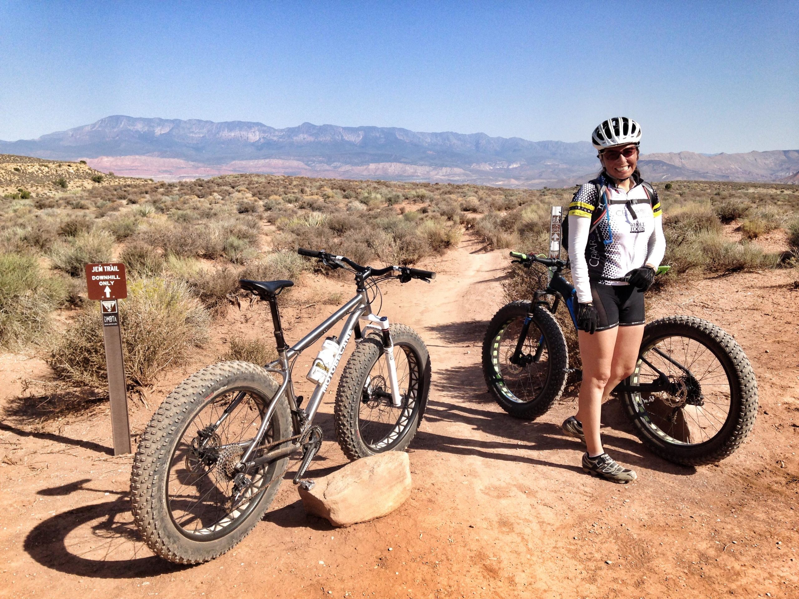 A female mountain biker stands next to two fat-tire bikes on a dirt trail, with a trail sign indicating "Jeep Trail" and "Downhill Only." The background features scenic mountains and clear blue skies. The area is surrounded by sparse desert vegetation. J.E.M. Trail mountain bike trail.