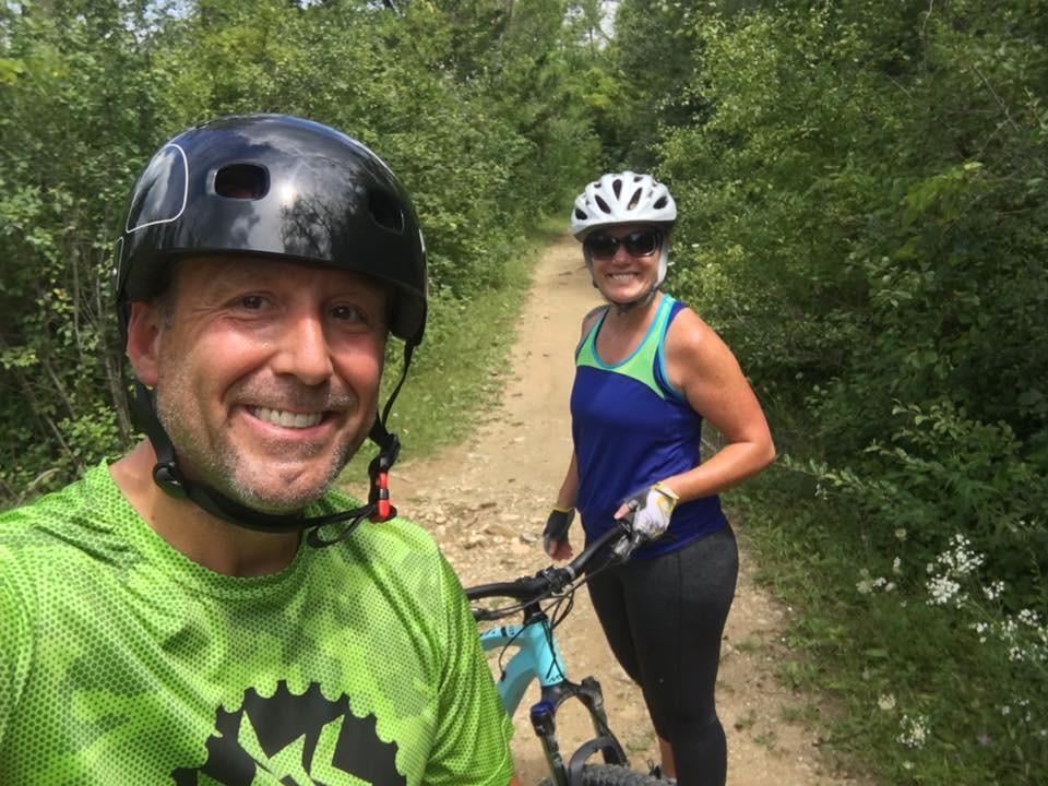 A smiling man and woman pose for a selfie on a dirt bike trail, surrounded by lush greenery. The man wears a black helmet and a green athletic shirt, while the woman, also in a helmet, is dressed in a blue tank top. Both appear excited and active, with mountain bikes beside them on the path. Saw Wee Kee Park mountain bike trail.