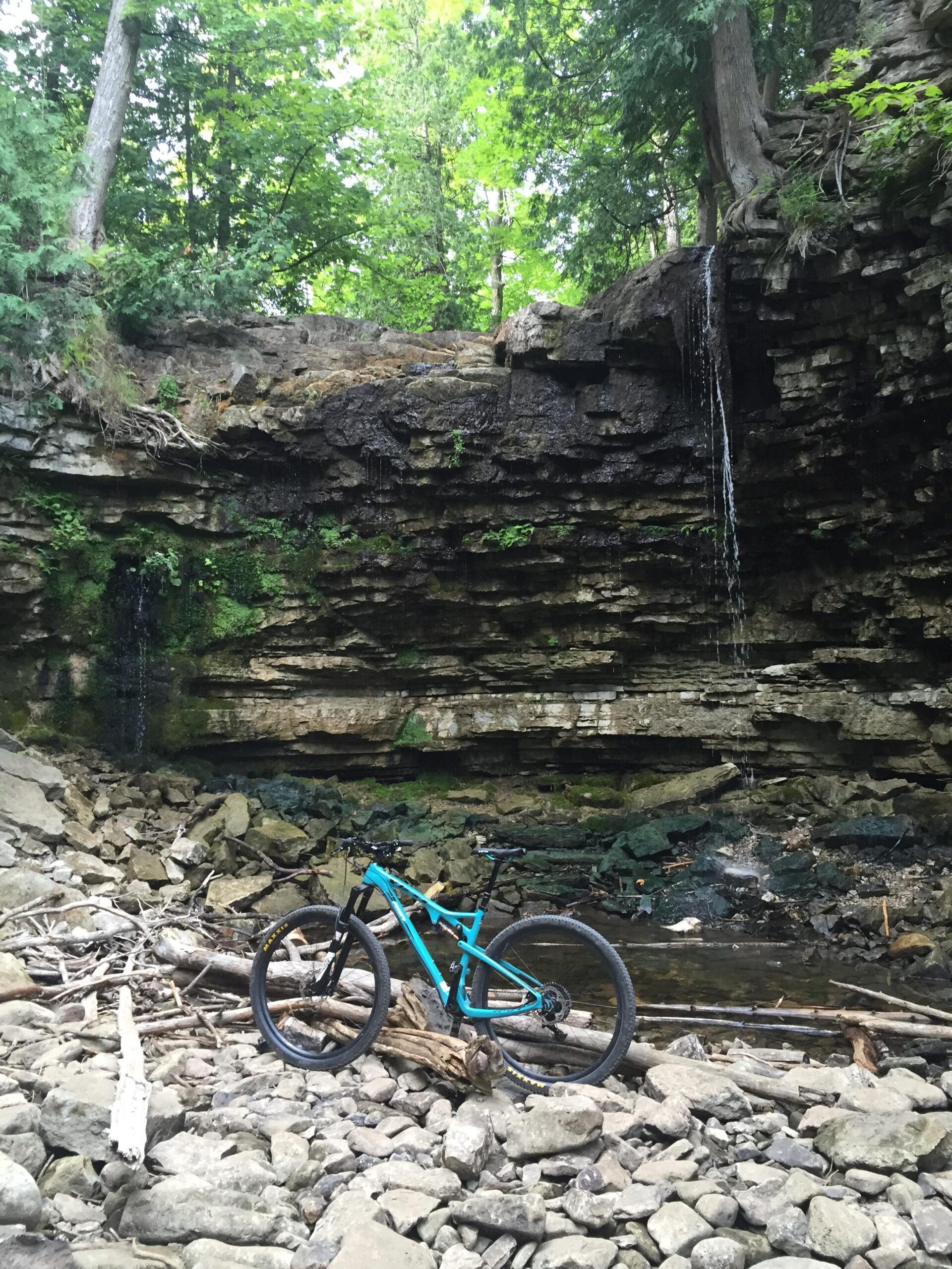 Yeti ASRc: A blue mountain bike rests on a rocky shore near a small waterfall and a wooded area. Lush green trees surround the scene, with water cascading gently down the moss-covered rock face in the background.