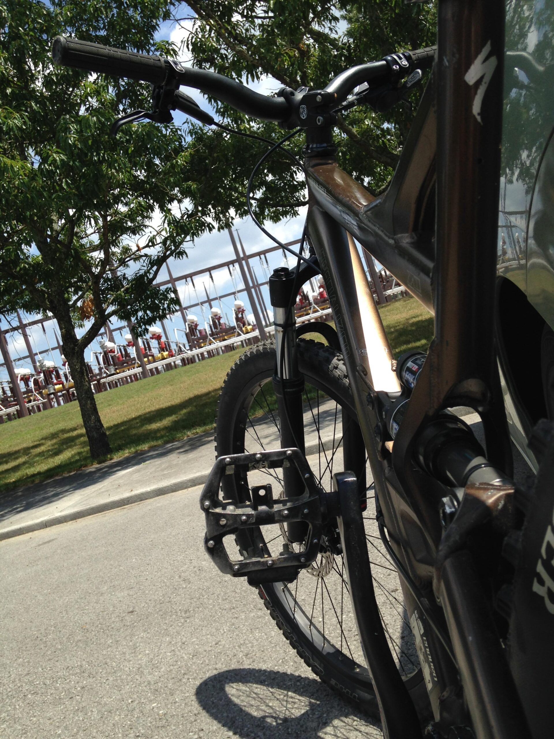 Specialized Enduro Pro: A close-up view of a mountain bike parked on a paved path, with a focus on the handlebars and pedal. In the background, a grassy area with people seated can be seen, along with trees and a blue sky.
