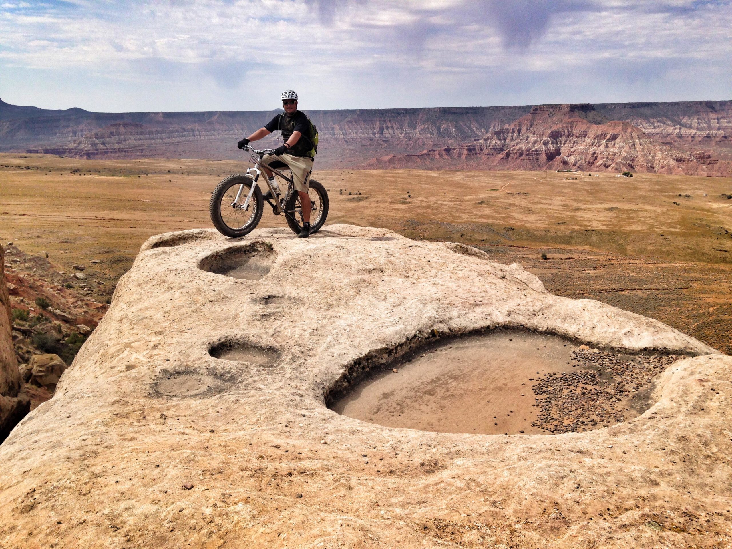 A mountain biker stands on a large, flat rock outcrop overlooking a vast canyon landscape. The scene features rugged terrain with distant hills and streaked cliffs under a partly cloudy sky. The biker is dressed in a helmet and casual cycling gear, and his bike rests on the rock beside him. The foreground shows unique rock formations with circular depressions, adding interest to the natural setting. Guacamole Mesa mountain bike trail.