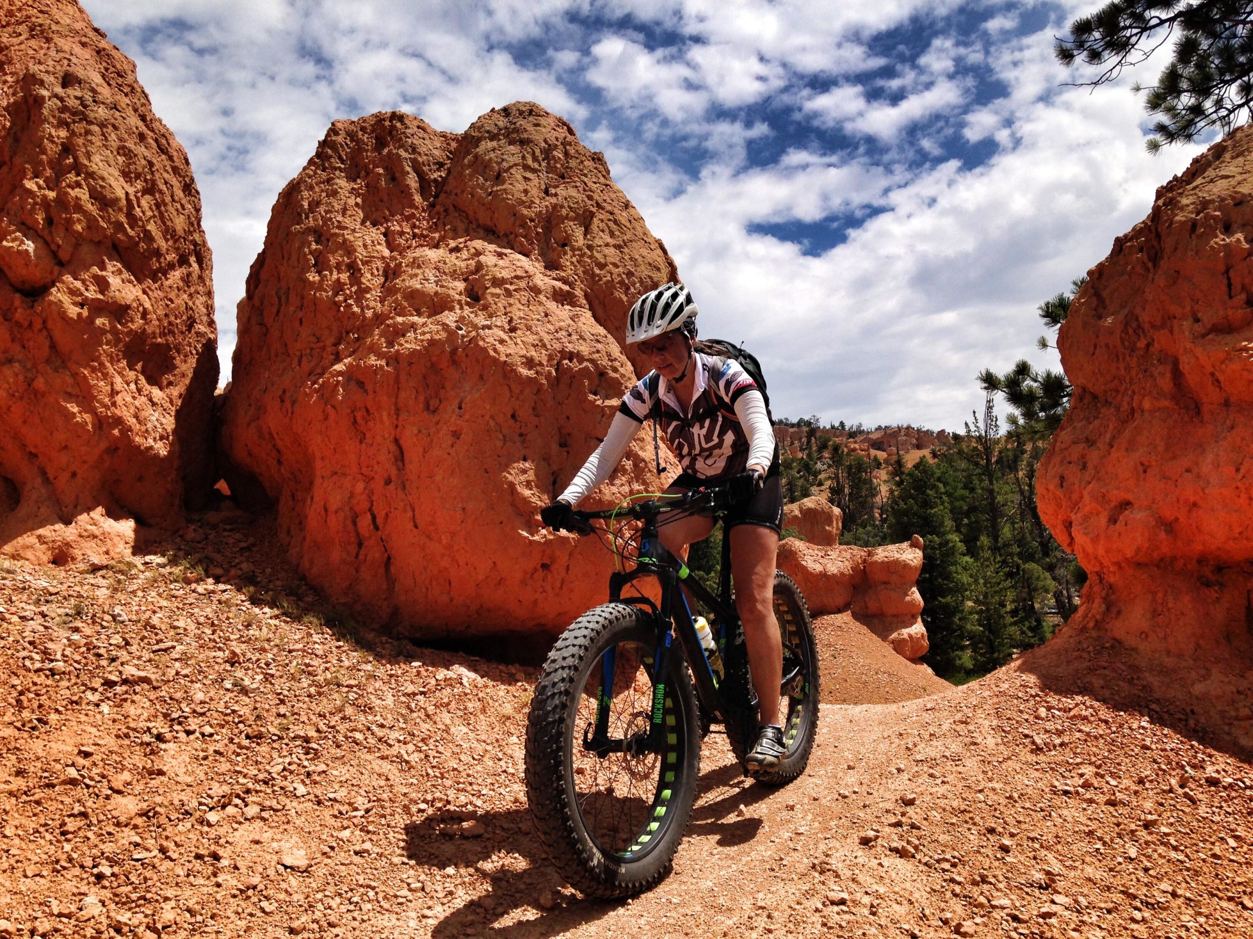 A mountain biker navigates a rocky trail surrounded by large orange rock formations under a partly cloudy sky. The rider, wearing a helmet and cycling gear, is focused on the path ahead. Pine trees are visible in the background, adding to the natural landscape. Thunder Mountain mountain bike trail.