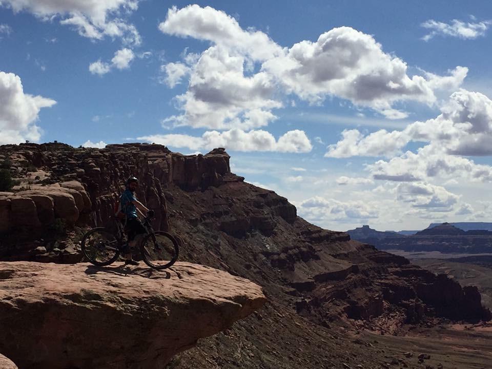 Giant Anthem X 29er 1: A mountain biker stands on a rocky ledge overlooking a vast canyon landscape, with dramatic cliffs and a blue sky filled with fluffy white clouds. The scene captures the sense of adventure and breathtaking scenery typical of outdoor biking trails.