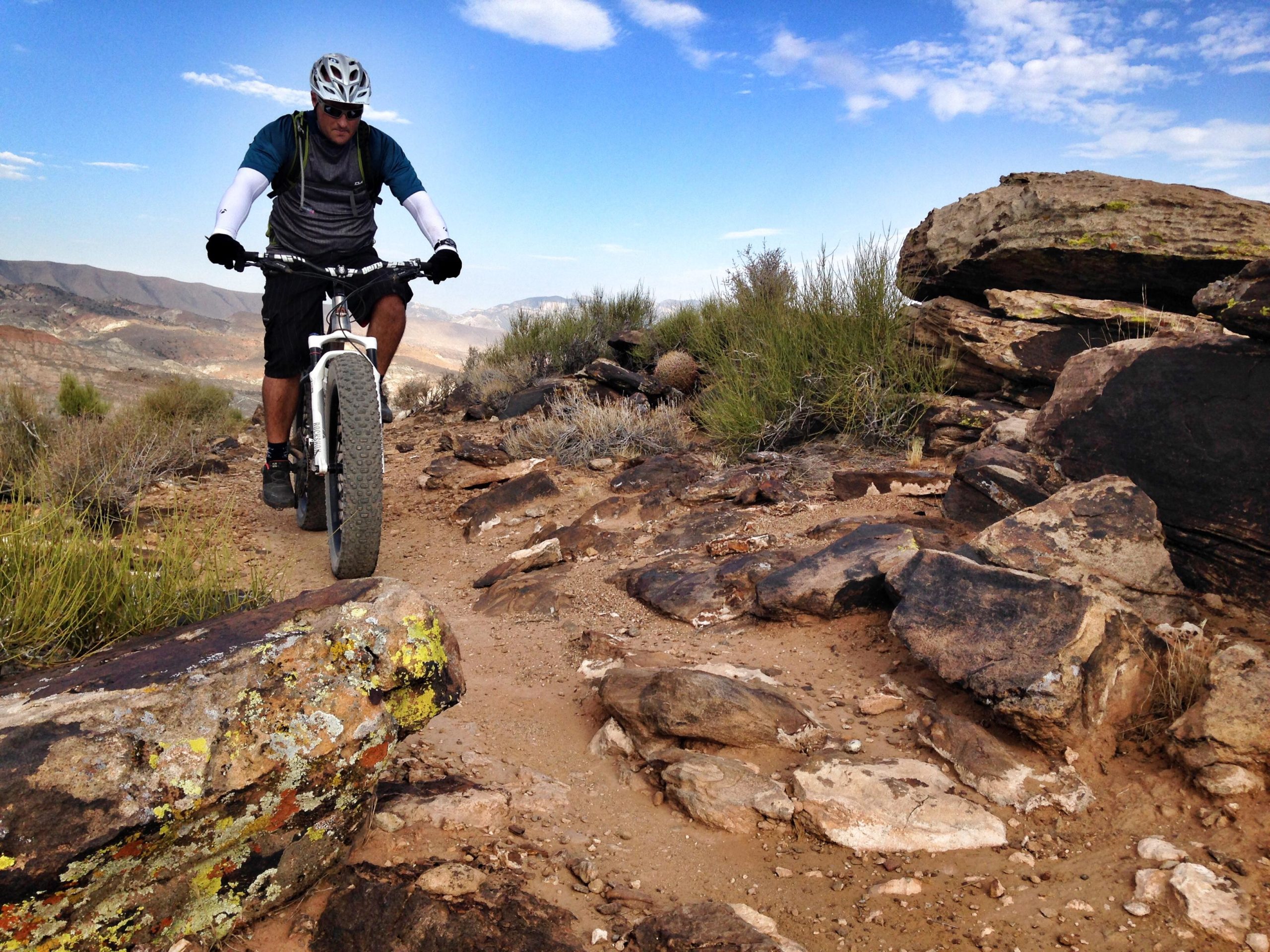 A cyclist navigating a rocky trail on a mountain bike in a scenic desert landscape, with shrubs and large boulders surrounding the path, under a clear blue sky. Barrel Roll mountain bike trail.
