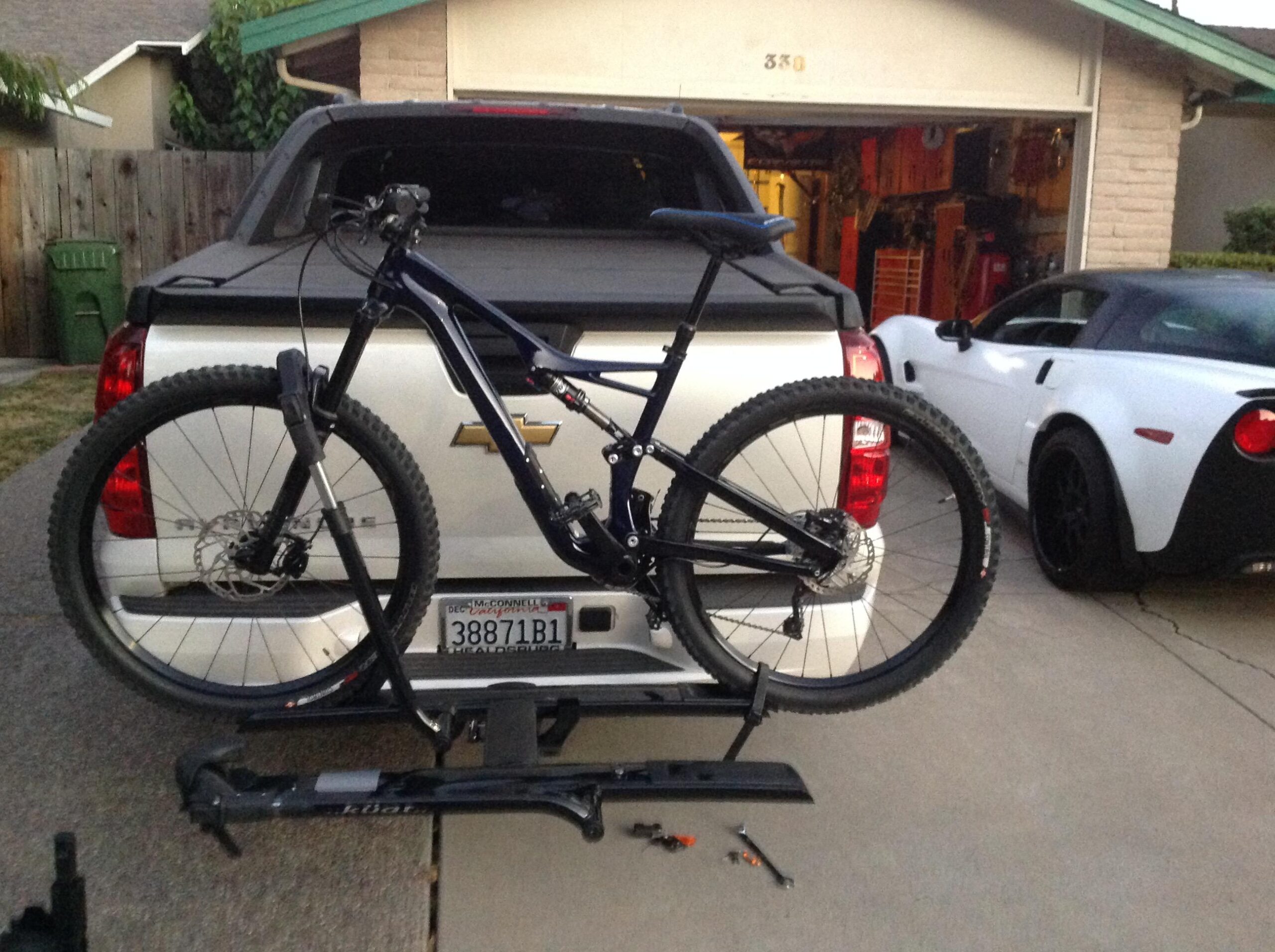Specialized Stumpjumper FSR comp carbon 650B: A mountain bike secured on the back of a Chevrolet pickup truck, parked in a driveway. A white sports car is visible to the right, and various tools are scattered on the ground nearby. The background features a garage with some equipment and a wooden fence.
