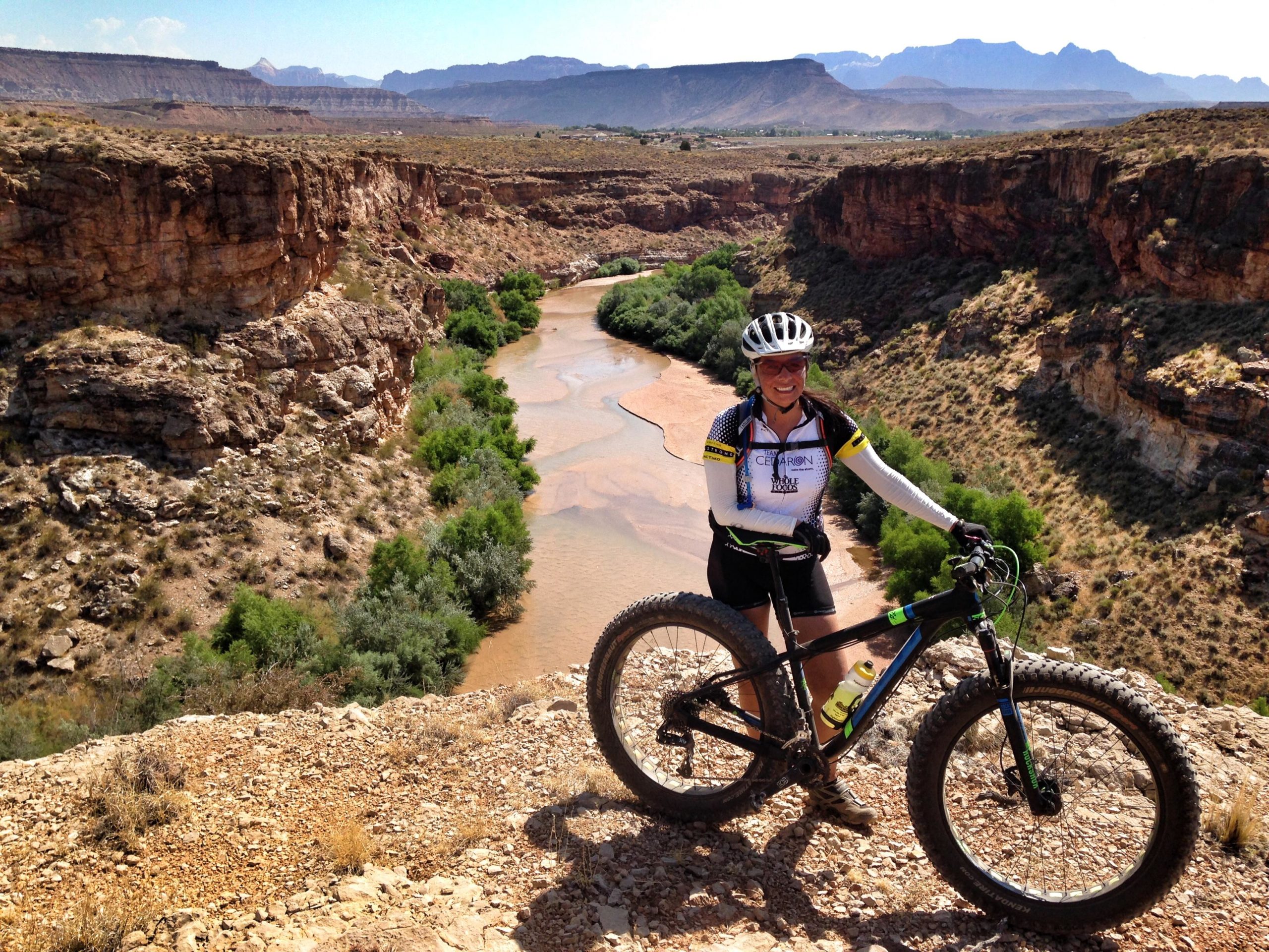 A person in athletic gear stands next to a mountain bike on a rocky overlook, with a river winding through a canyon below and mountains in the background. The scene is sunny, showcasing a rugged landscape with greenery along the riverbanks. J.E.M. Trail mountain bike trail.