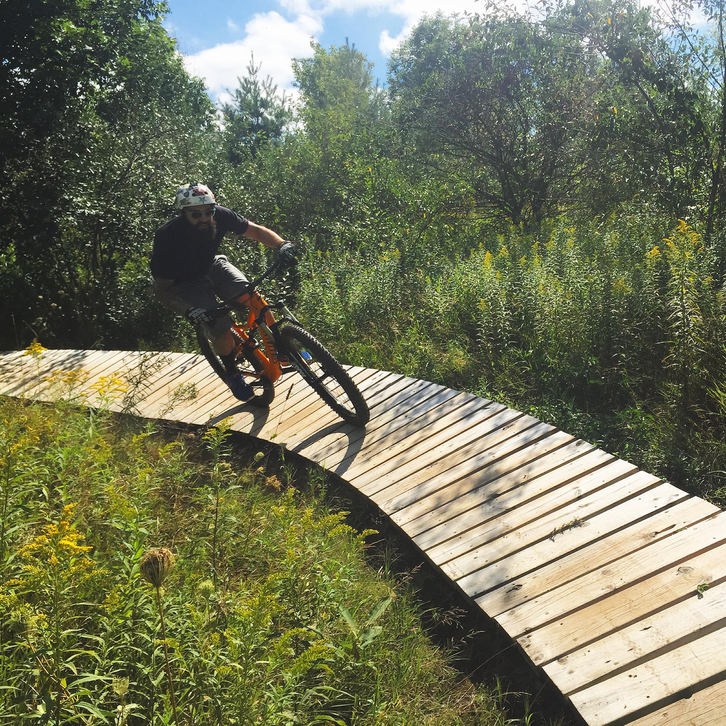 Specialized Stumpjumper FSR Comp 6Fattie: A person riding a mountain bike on a curved wooden path through a lush green landscape. The rider, wearing a helmet and protective gear, is navigating the trail surrounded by trees and tall grasses under a blue sky.