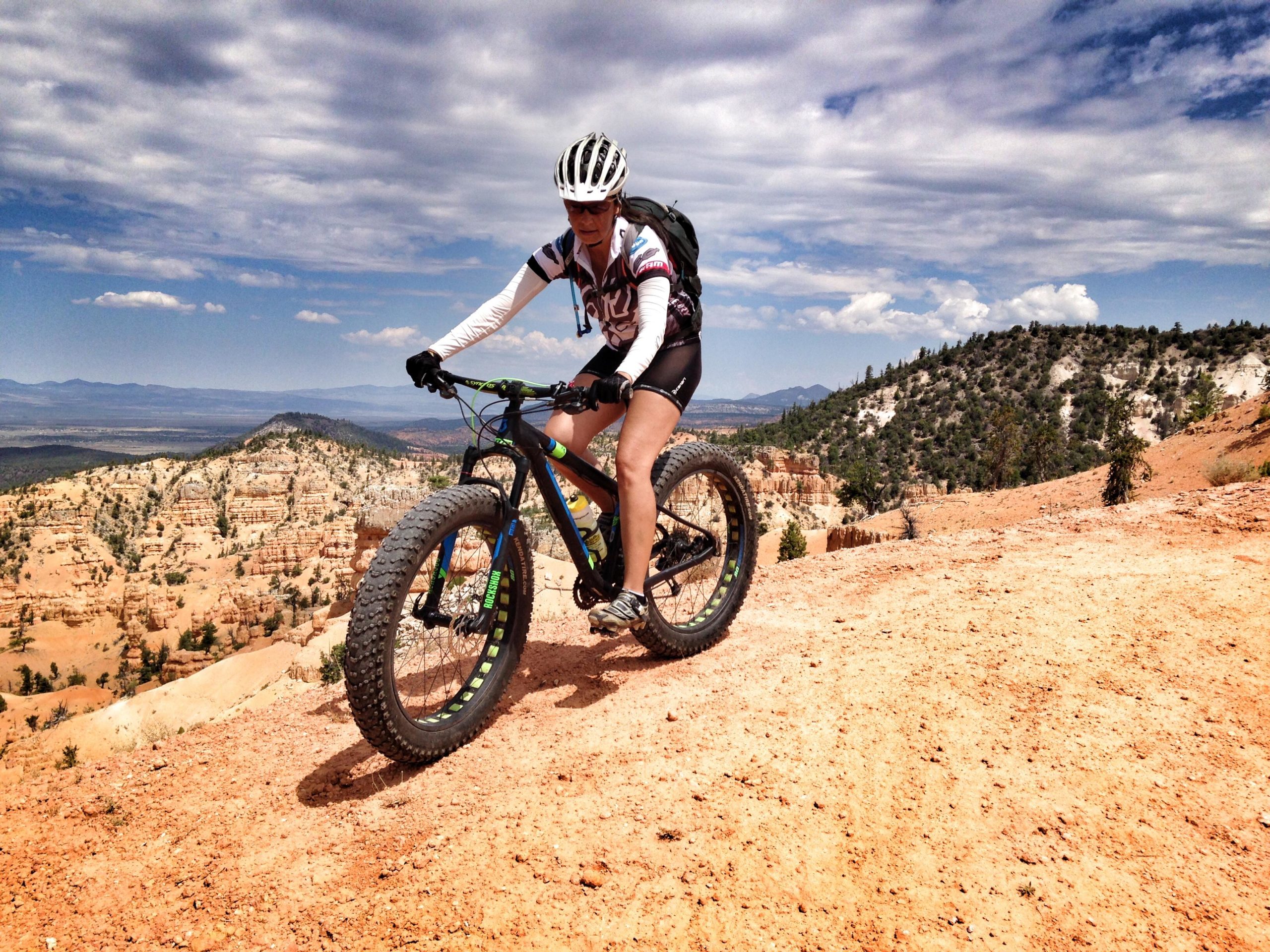 A cyclist riding a fat-tire mountain bike on a rocky dirt trail, surrounded by stunning desert landscapes and canyons with a blue sky and fluffy clouds in the background. Thunder Mountain mountain bike trail.