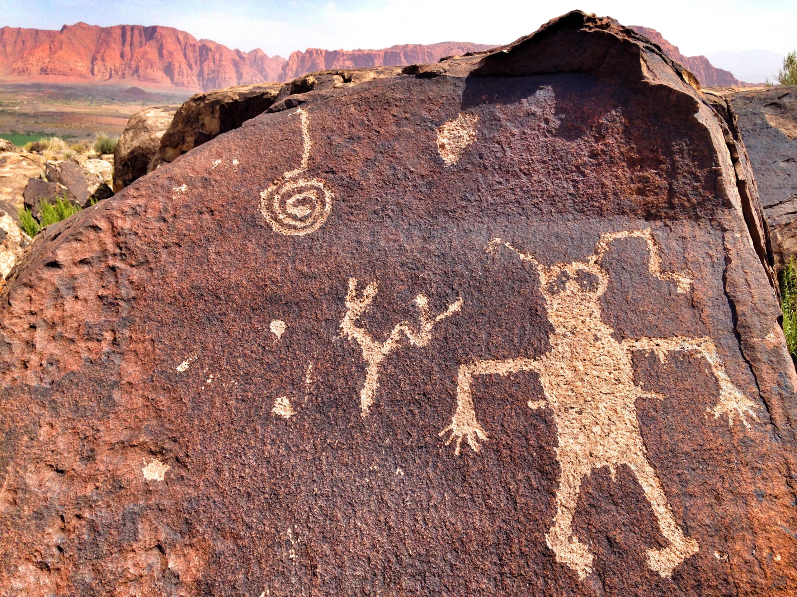 Petroglyphs carved into a large rock surface, depicting abstract figures and symbols, including a spiral, alongside a humanoid figure with outstretched arms, set against a backdrop of red mountains and clear skies. Barrel Roll mountain bike trail.