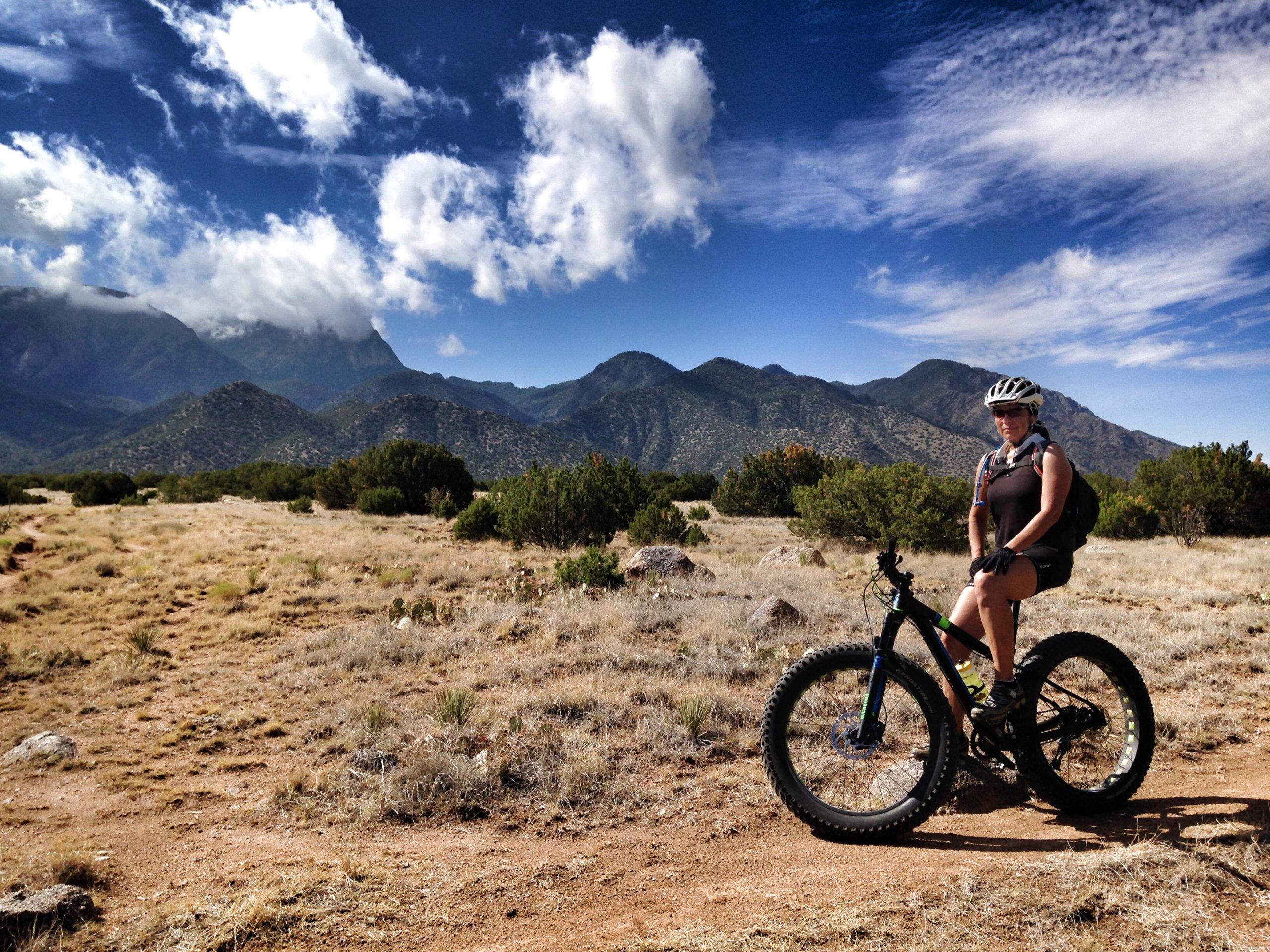 A cyclist on a fat bike is seated on a dirt path in a mountainous landscape. The background features rolling hills and mountains under a partly cloudy blue sky, while the foreground has sparse grass and small bushes. The cyclist is wearing a helmet and cycling gear, with a backpack visible. Placitas Trails mountain bike trail.