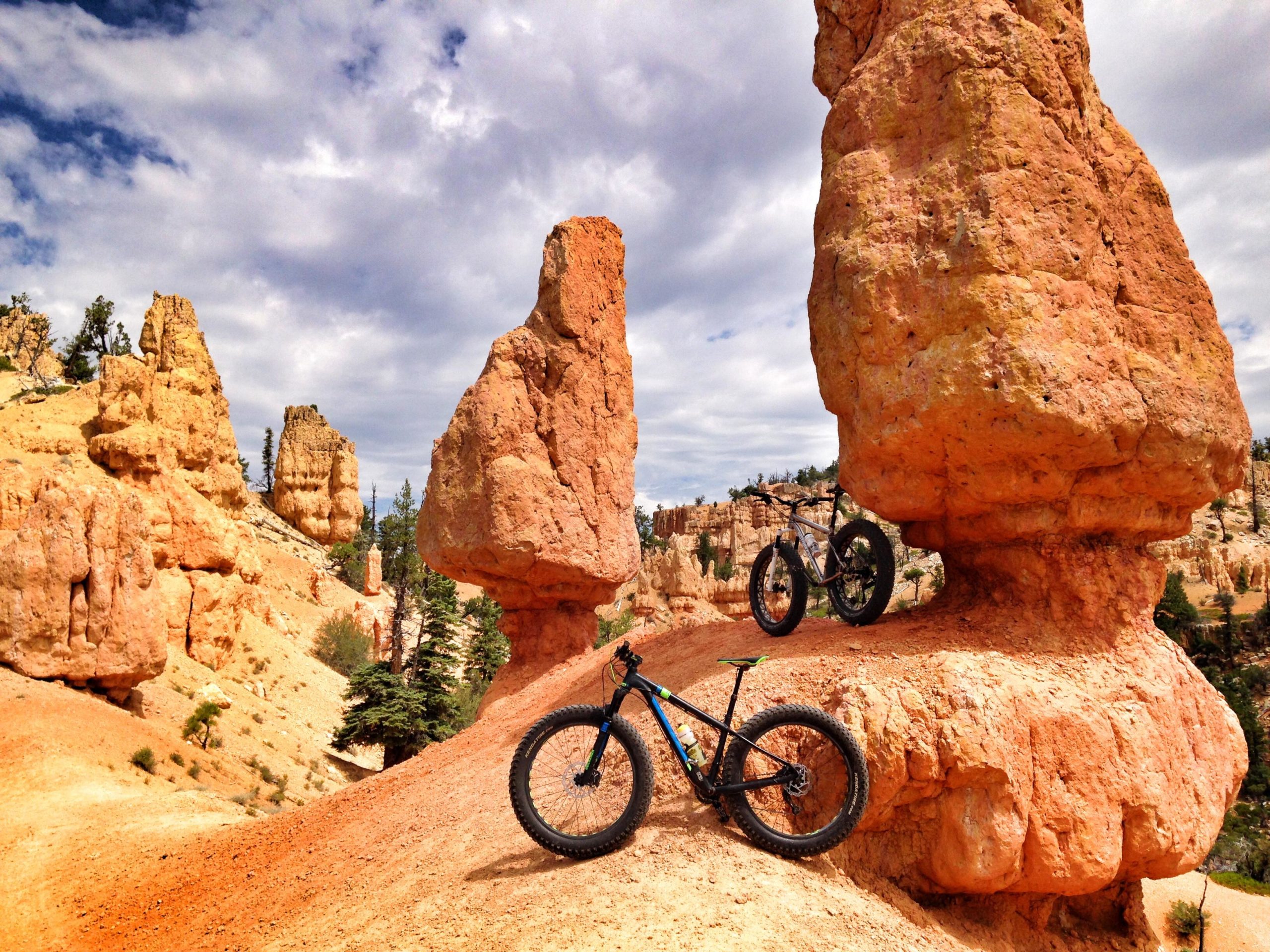 Two mountain bikes are positioned on a rocky, orange landscape with unique rock formations in the background. The setting features towering hoodoos under a partly cloudy sky, surrounded by forested areas. The image captures the essence of outdoor adventure and mountain biking in a scenic natural environment. Thunder Mountain mountain bike trail.