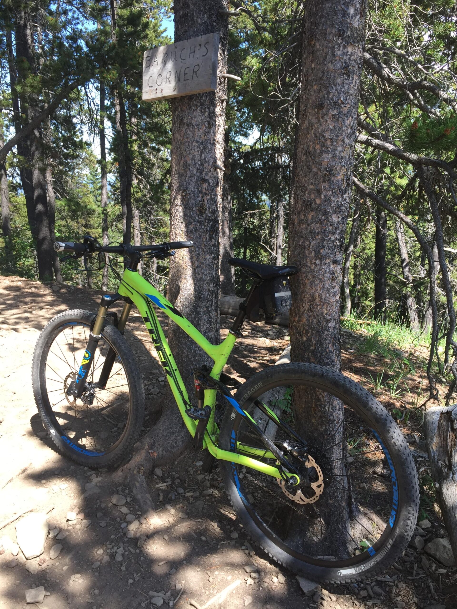 Trek Fuel EX 9 29: A bright green mountain bike leaning against a tree with a wooden sign reading "Barduch's Corner" in a forested area. Sunlight filters through the trees, illuminating the dirt path and surrounding vegetation.
