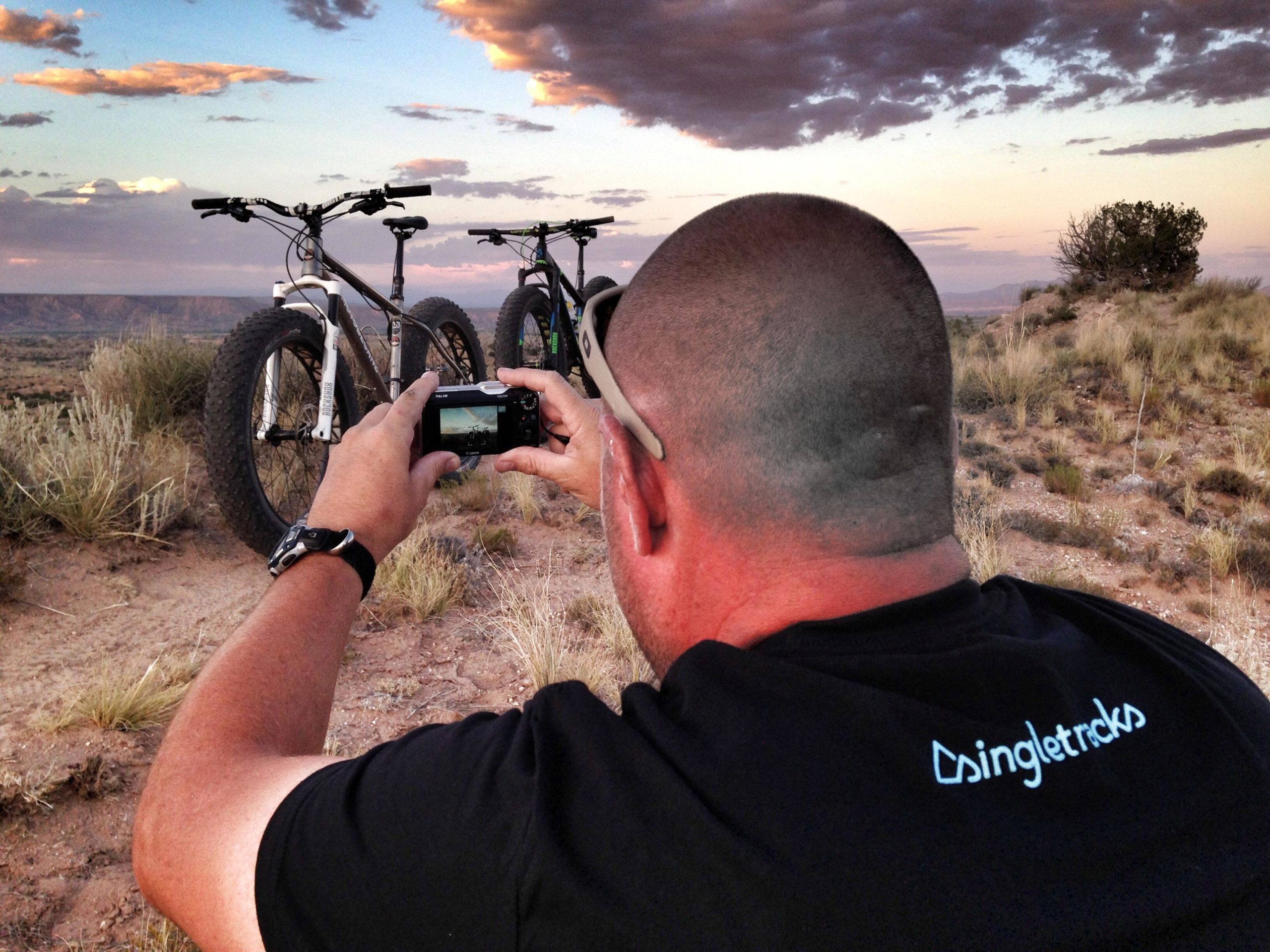 A person wearing a black shirt with the text "singletracks" is taking a photo with a camera. Two fat tire bicycles are positioned in the foreground against a scenic backdrop of a sunset sky and a distant landscape filled with shrubs and hills. The lighting creates a warm atmosphere, highlighting the details of the bicycles and the surrounding nature. Mariposa Fat Bike Trails mountain bike trail.