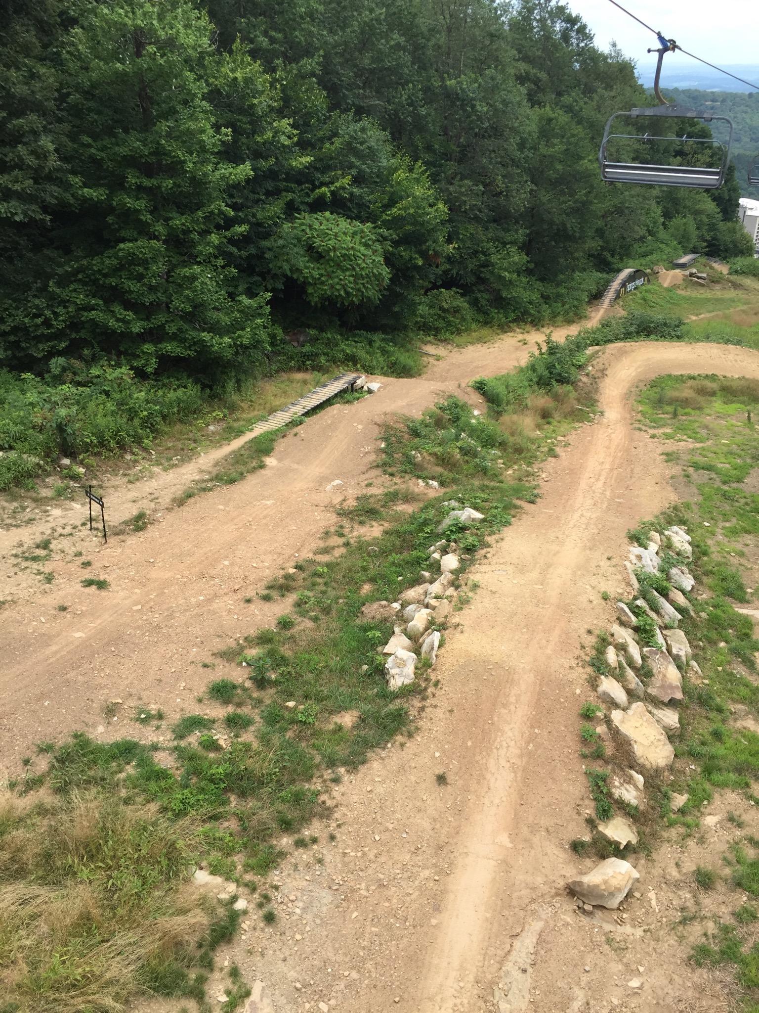 A view from a chairlift overlooking a dirt trail network, with winding paths and a wooden bridge. Surrounding the trails are patches of grass, rocks, and dense green trees, creating a natural landscape. Seven Springs mountain bike trail.