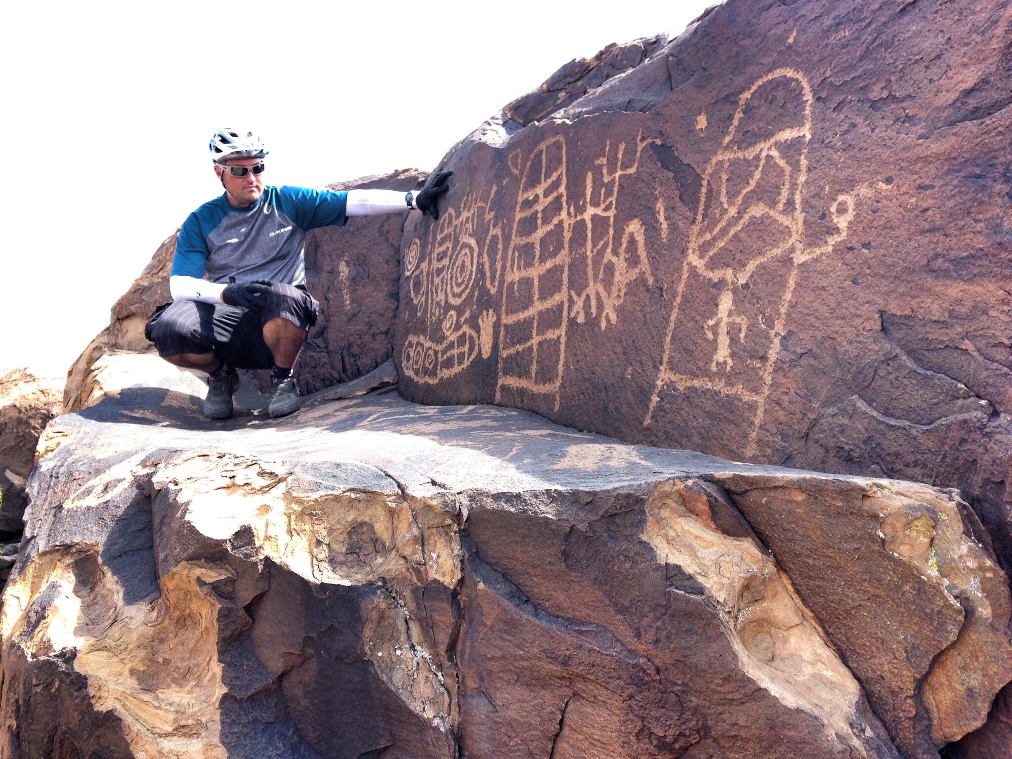 A person in athletic gear poses near ancient petroglyphs etched into a large rock surface. The petroglyphs depict various abstract designs and figures, showcasing the area's historical significance. The individual is wearing a helmet and gloves, indicating they're engaged in an outdoor activity, possibly biking or hiking. The scene is set in a bright, open environment. Barrel Roll mountain bike trail.