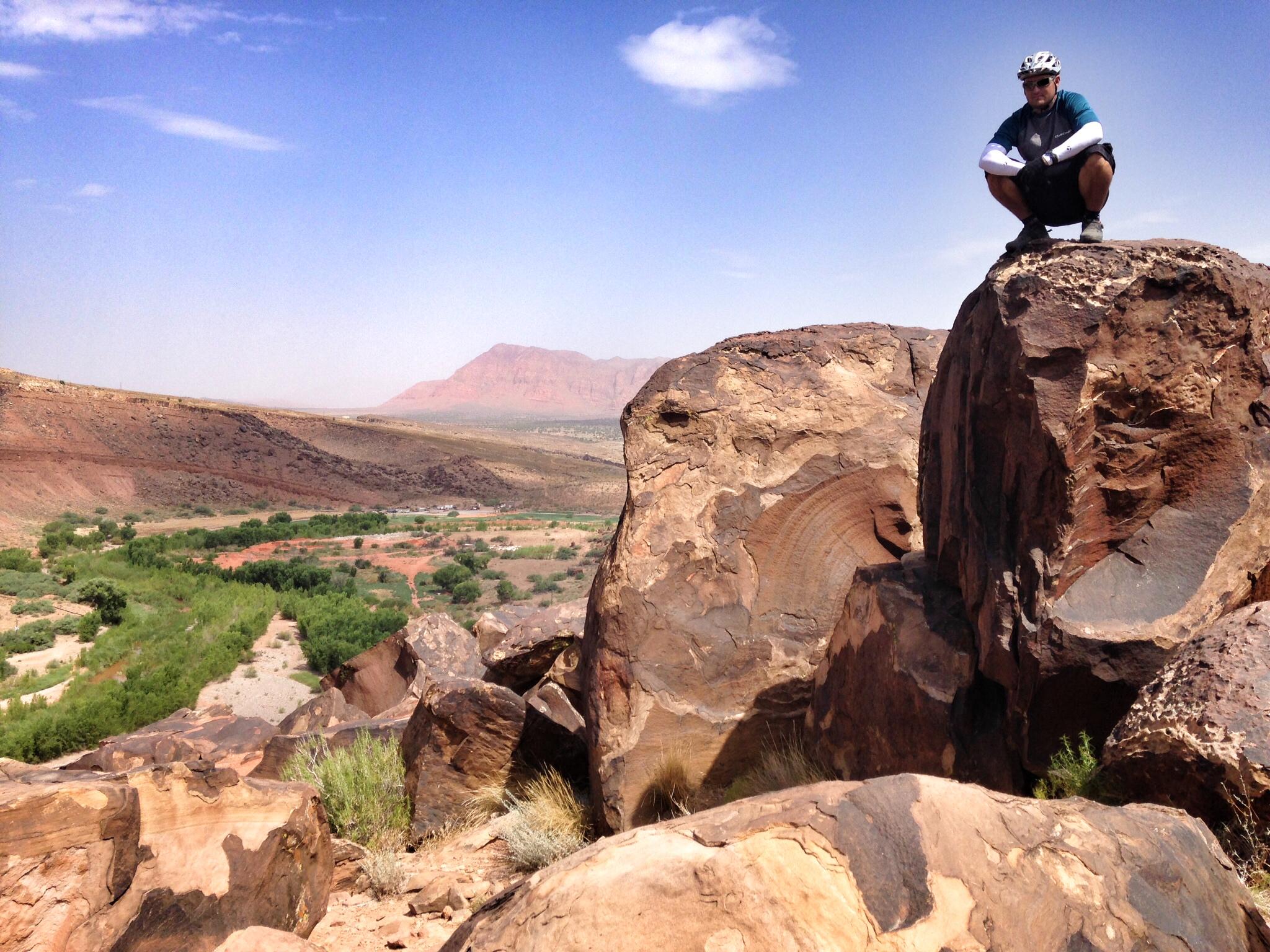 A person wearing a helmet and cycling gear is crouched on a large rock overlooking a vast desert landscape with hills and a river below. The sky is clear with a few clouds, and a distant mountain range is visible in the background. The scene captures the rugged natural beauty of the area. Barrel Roll mountain bike trail.