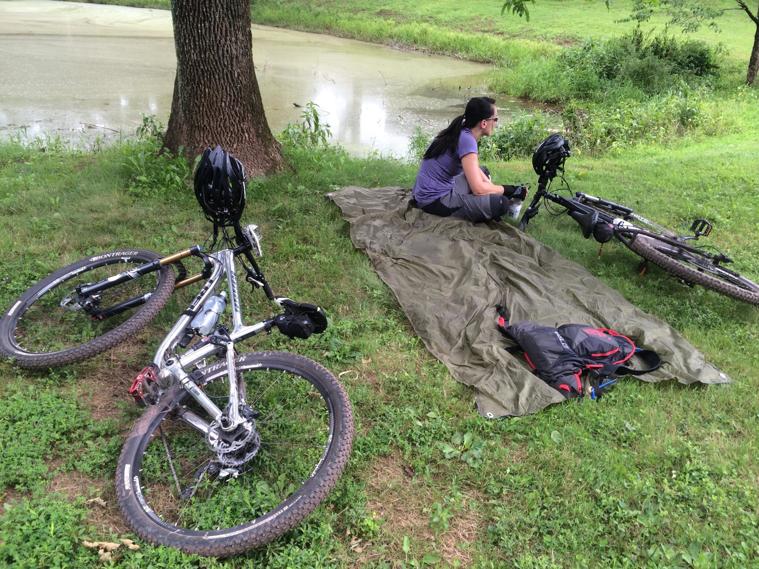 A person sitting on a green tarp by a pond, wearing a gray t-shirt and cycling gear, with two mountain bikes lying on the grass nearby. One bike has a black helmet resting on it, while a backpack in dark colors is placed next to the bikes. The surrounding area features lush grass and trees. Six Mile Run mountain bike trail.