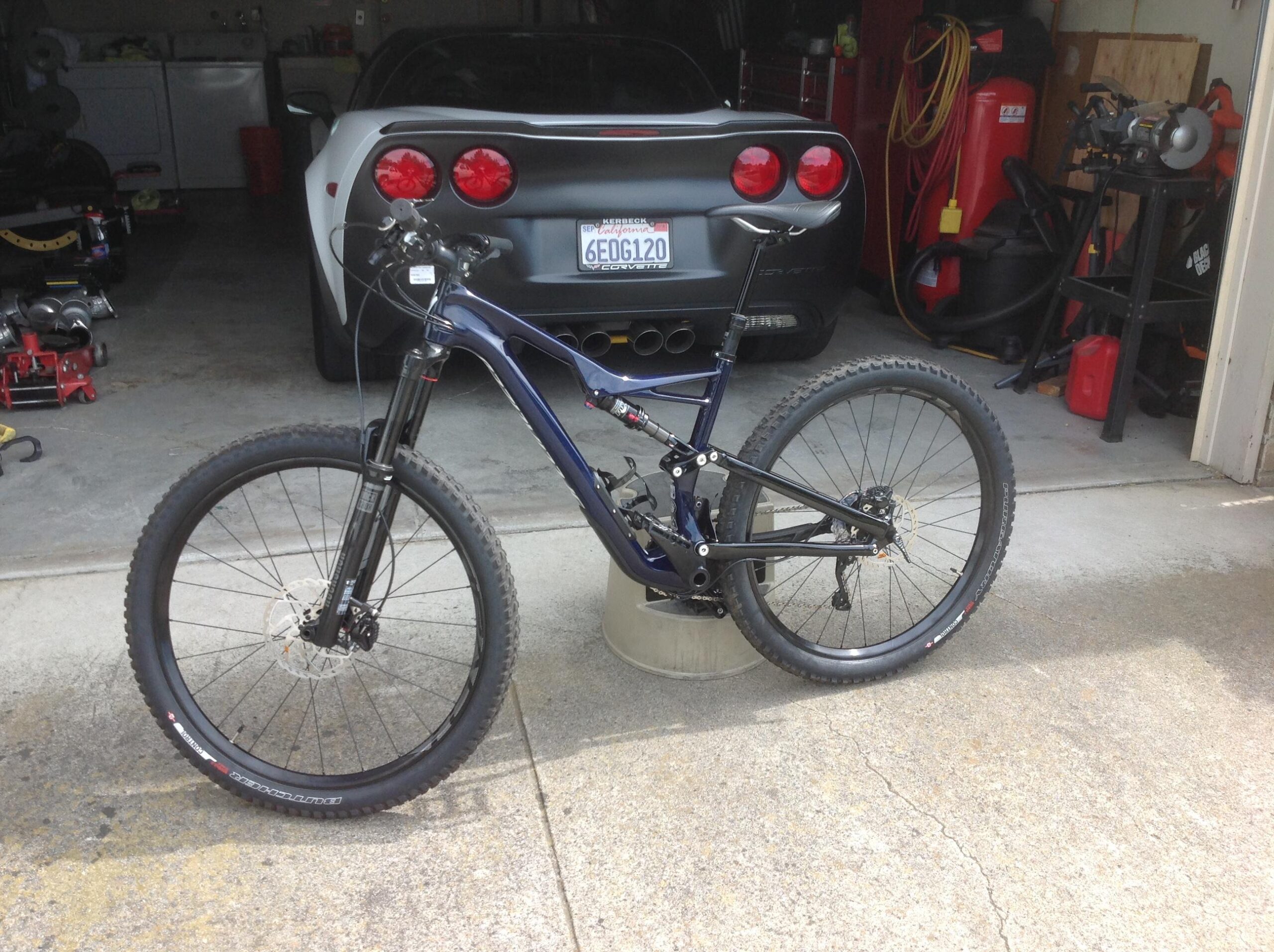 Specialized Stumpjumper FSR comp carbon 650B: A blue mountain bike standing on a concrete surface in a garage, with a black sports car visible in the background. Various tools and equipment are arranged around the garage setting.