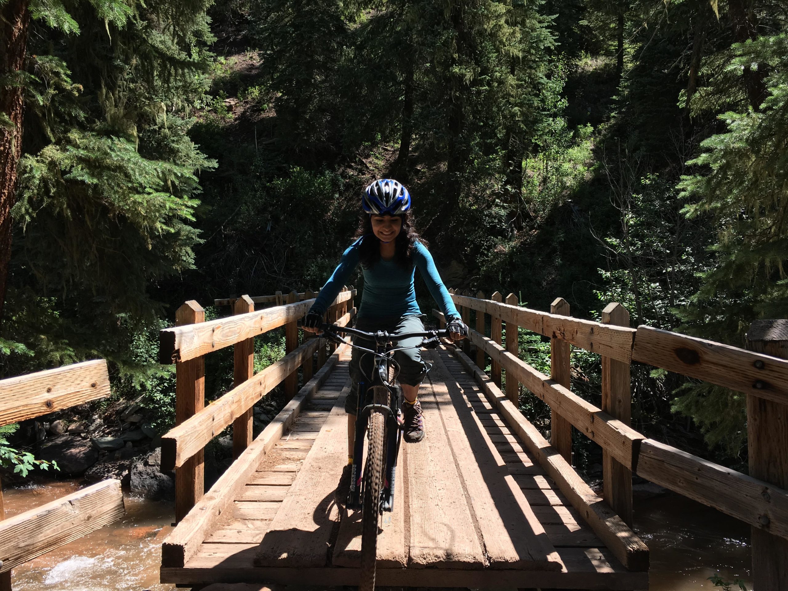 A person wearing a blue helmet and a long-sleeve shirt rides a mountain bike across a wooden bridge surrounded by lush greenery and trees. Sunlight filters through the foliage, creating a bright and vibrant atmosphere. Hermosa Creek Trail mountain bike trail.