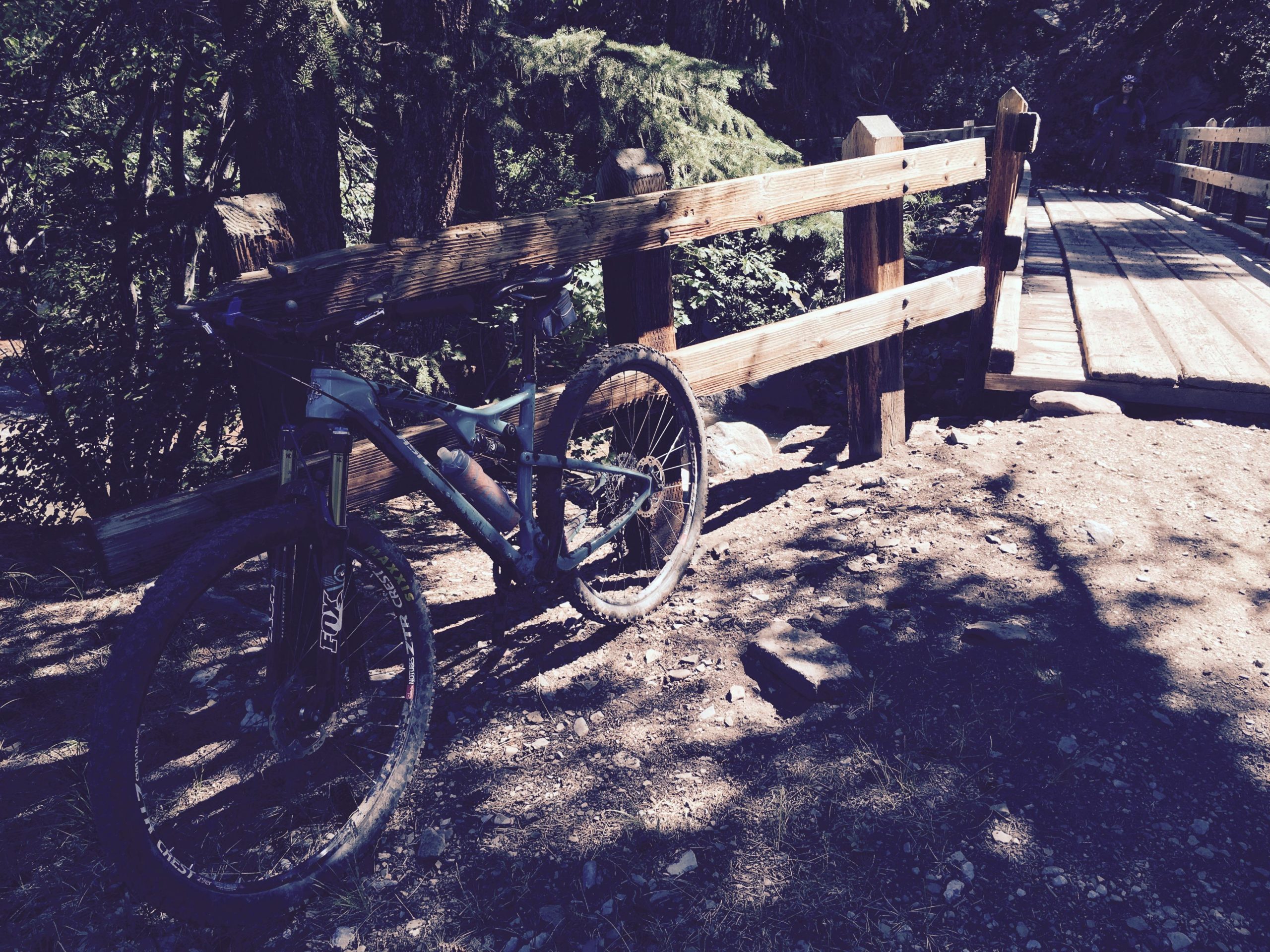 A mountain bike resting against a wooden railing near a bridge in a forested area. Sunlight filters through the trees, casting dappled shadows on the ground. The bridge spans a small creek, surrounded by greenery. Hermosa Creek Trail mountain bike trail.