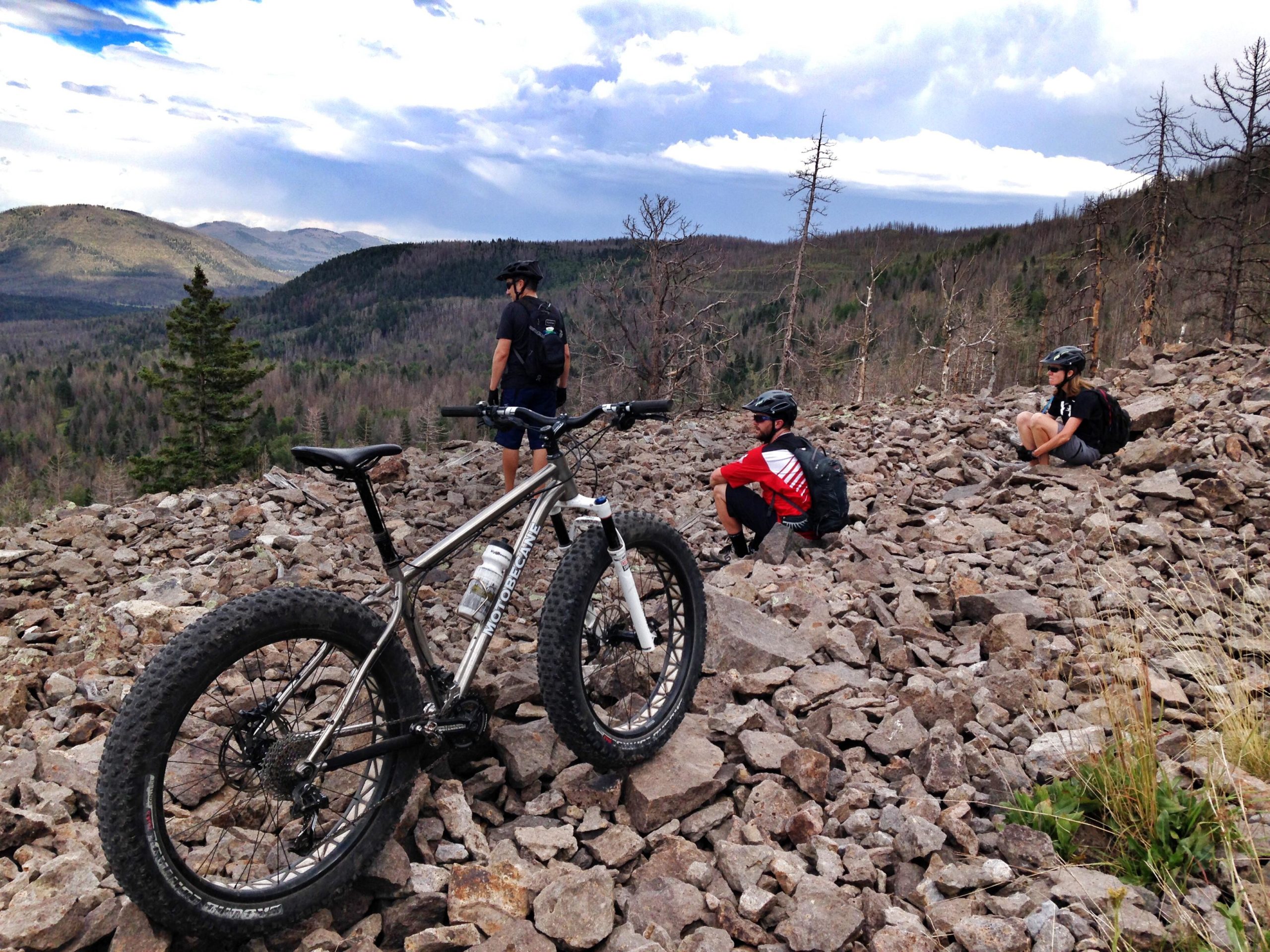 Three mountain bikers resting on a rocky hillside, with one rider standing and looking out over a scenic landscape of mountains and valleys. A fat tire bike is in the foreground, and the background features sparse trees and a cloudy sky. Valles Caldera Trails mountain bike trail.