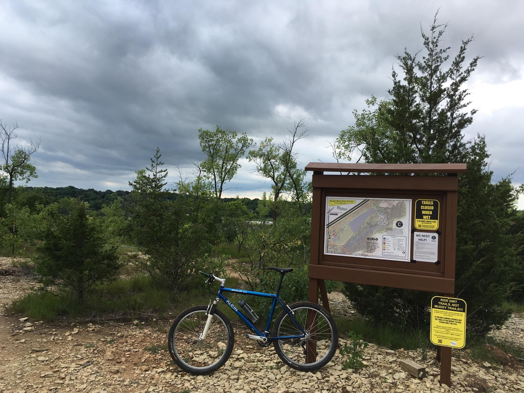 Klein Attitude Race: A mountain bike rests beside an informational sign at a trailhead, with a detailed map and trail guidelines. The background features a mix of green trees under a cloudy sky, indicating a natural outdoor setting.