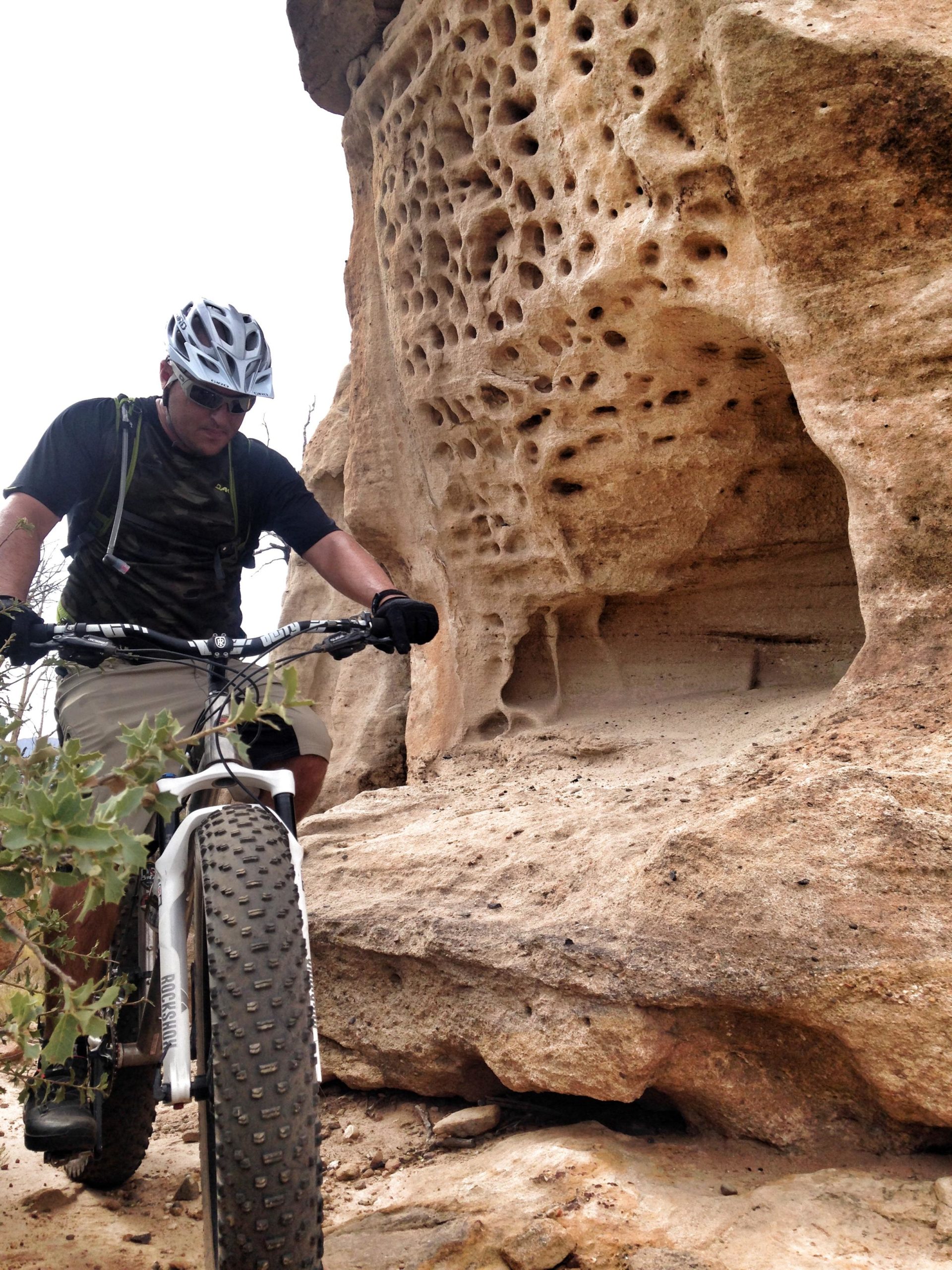 A person riding a mountain bike on a rocky trail, leaning slightly forward while navigating past a textured rock formation with holes and indentations. The surroundings feature sparse vegetation, and the cyclist is wearing a helmet and sunglasses, dressed in a short-sleeve shirt and shorts, indicative of an outdoor adventure in a natural environment. Guacamole Mesa mountain bike trail.