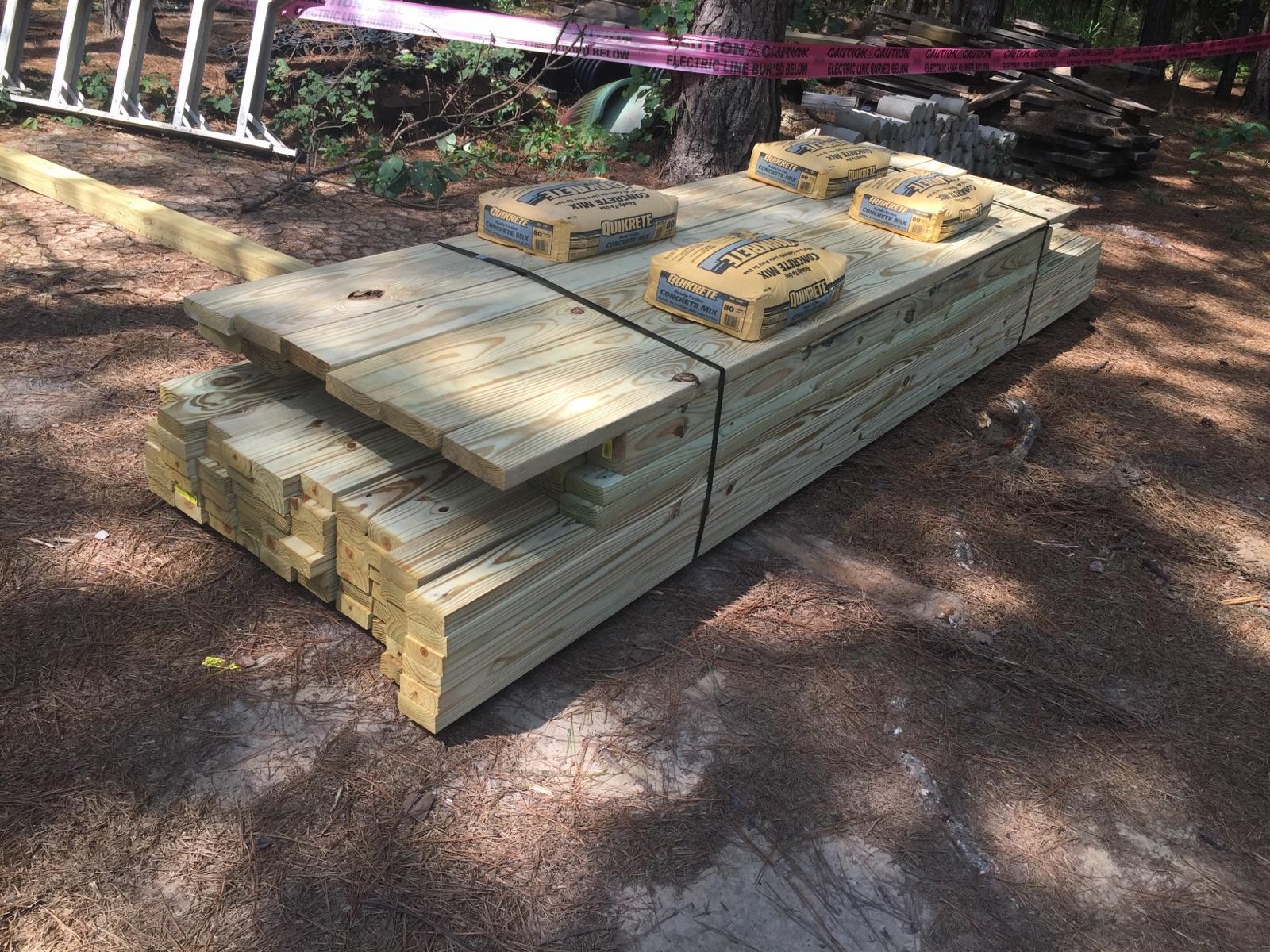A stack of wooden lumber boards and construction bags is resting on the ground in a wooded area. The lumber is neatly arranged, with several long planks on top and shorter pieces underneath. Construction bags are placed on top of the lumber for added weight. In the background, a ladder and other construction materials can be seen, along with a caution tape indicating a work zone. Mt. Zion Bike Trails mountain bike trail.
