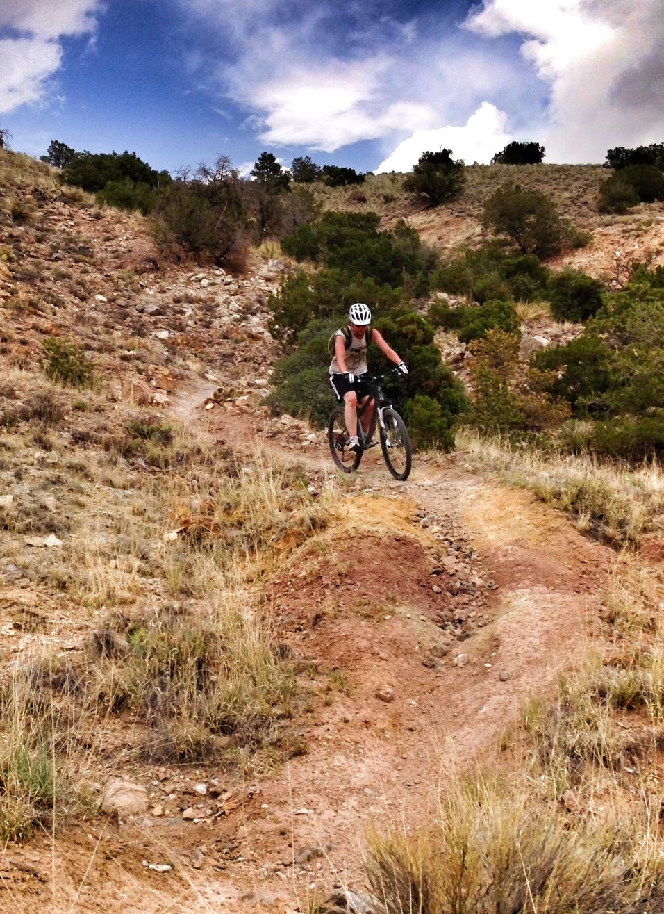 A person riding a mountain bike down a dirt trail surrounded by rocky terrain and sparse vegetation, under a partly cloudy sky. The cyclist is wearing a helmet and casual attire, navigating a slope that features a mix of grassy and earthy patches. Chalk Dust mountain bike trail.