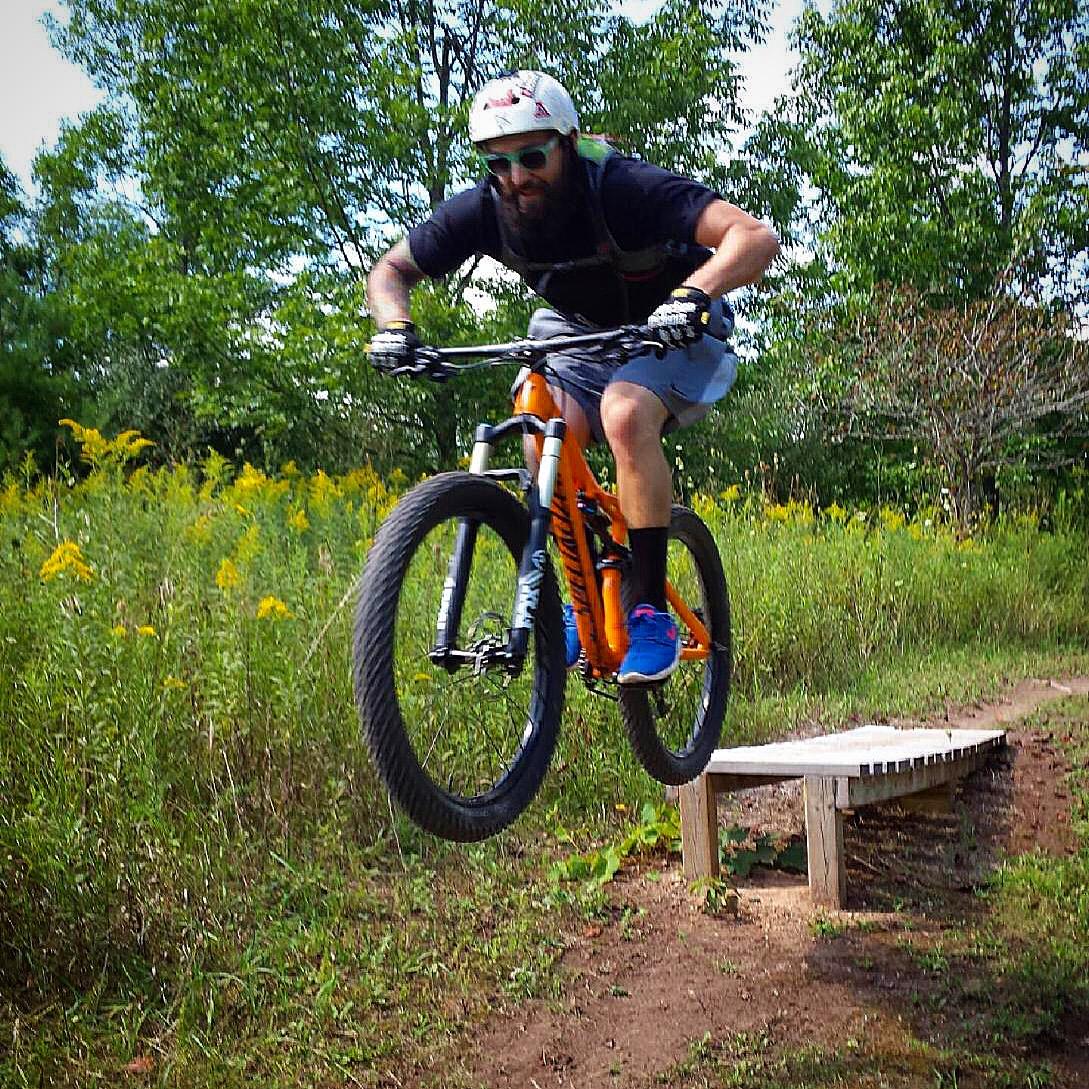 Specialized Stumpjumper FSR Comp 6Fattie: A mountain biker in a black shirt and helmet performs a jump over a wooden ramp on a dirt trail surrounded by tall green grass and trees. The cyclist’s orange bike is airborne, showcasing an exciting moment in a nature setting.