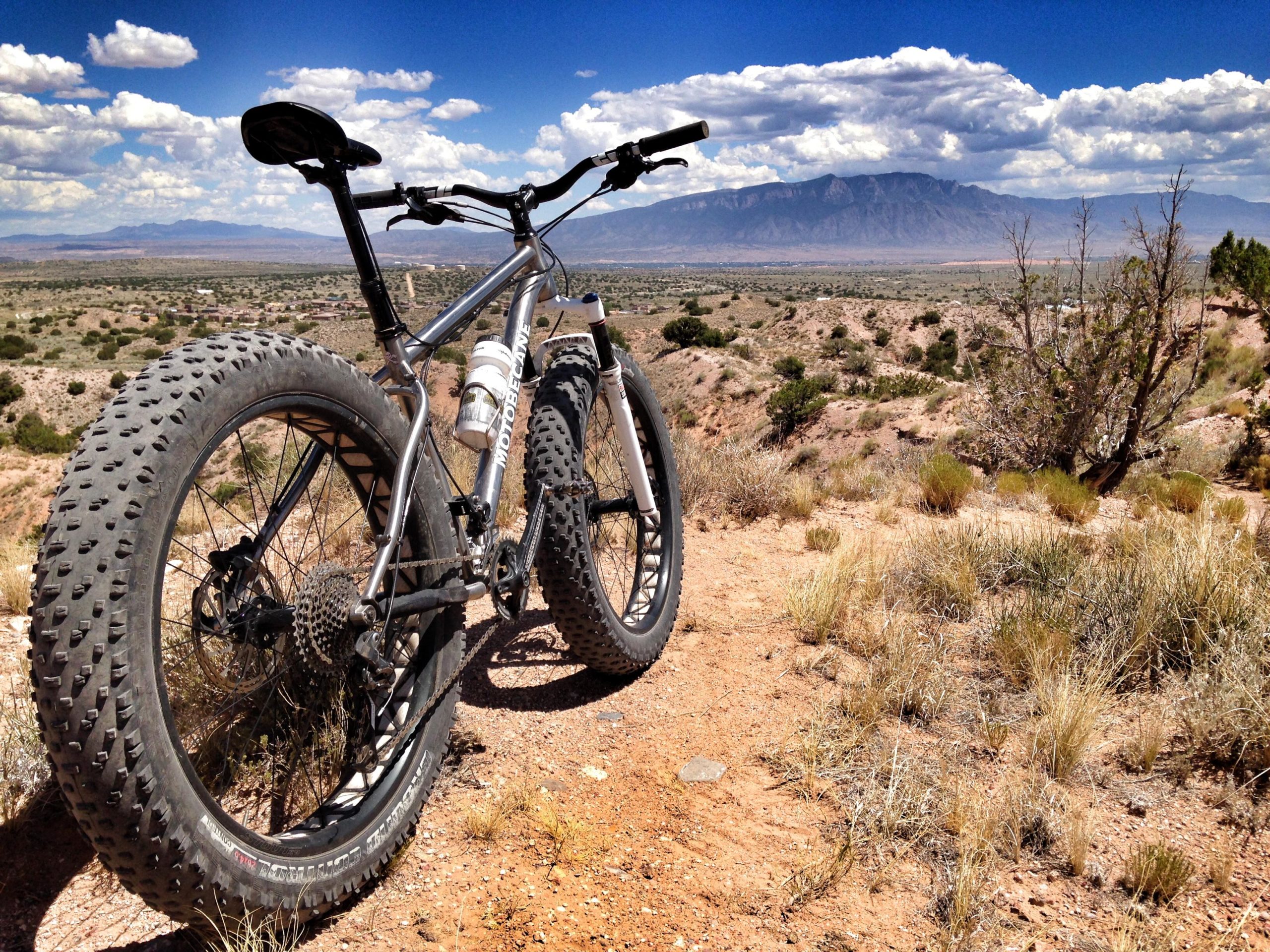 A fat tire bike positioned on a rocky terrain, overlooking a vast landscape with distant mountains and a blue sky filled with clouds. The bike's large tires and sturdy frame suggest it's designed for rugged trails. Shrubs and grasses grow around the bike, enhancing the natural scenery. Rim Trail mountain bike trail.