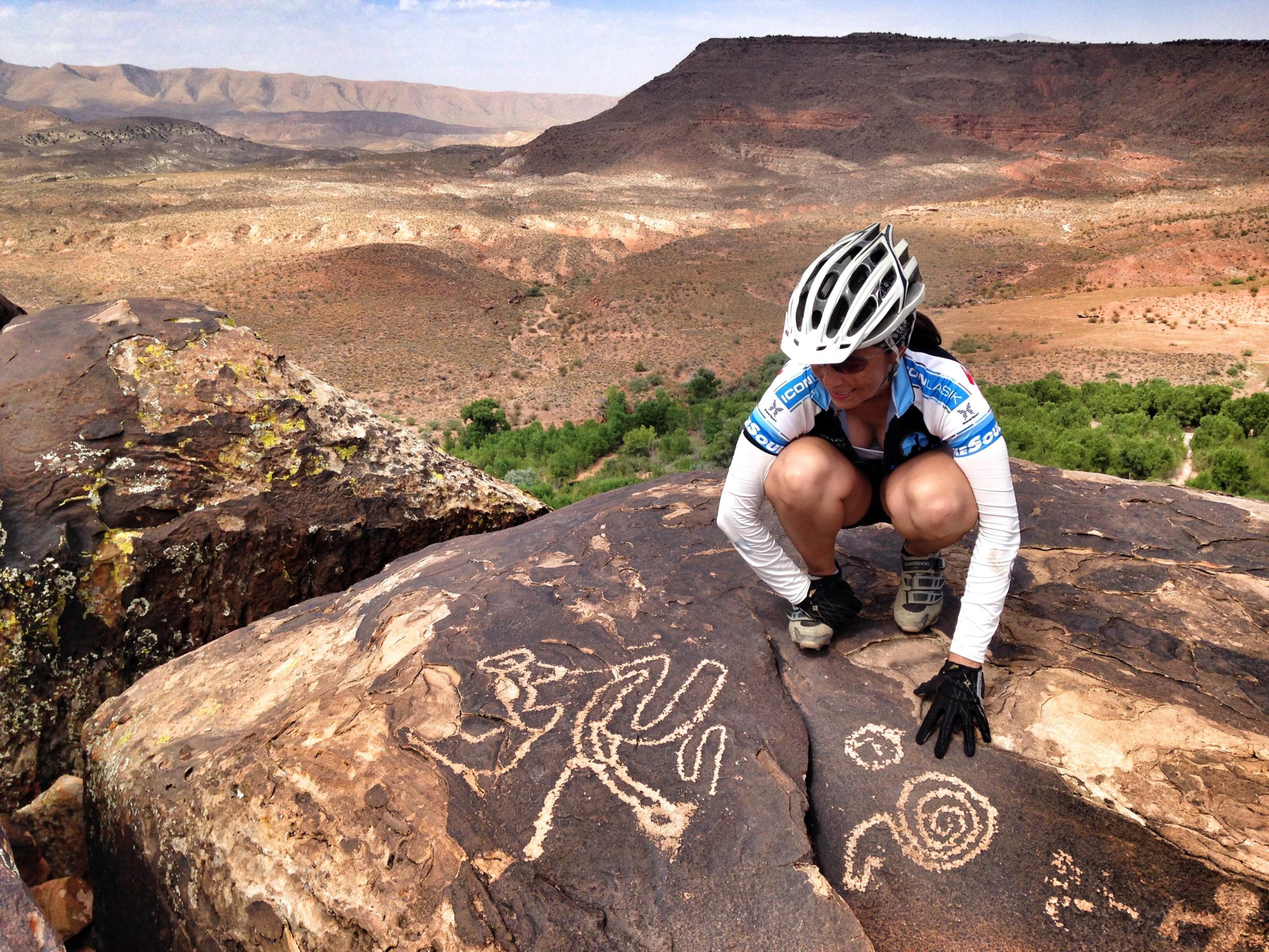 A woman in cycling gear crouches on a rocky surface, examining ancient petroglyphs in a desert landscape with distant mountains and greenery below. Barrel Roll mountain bike trail.