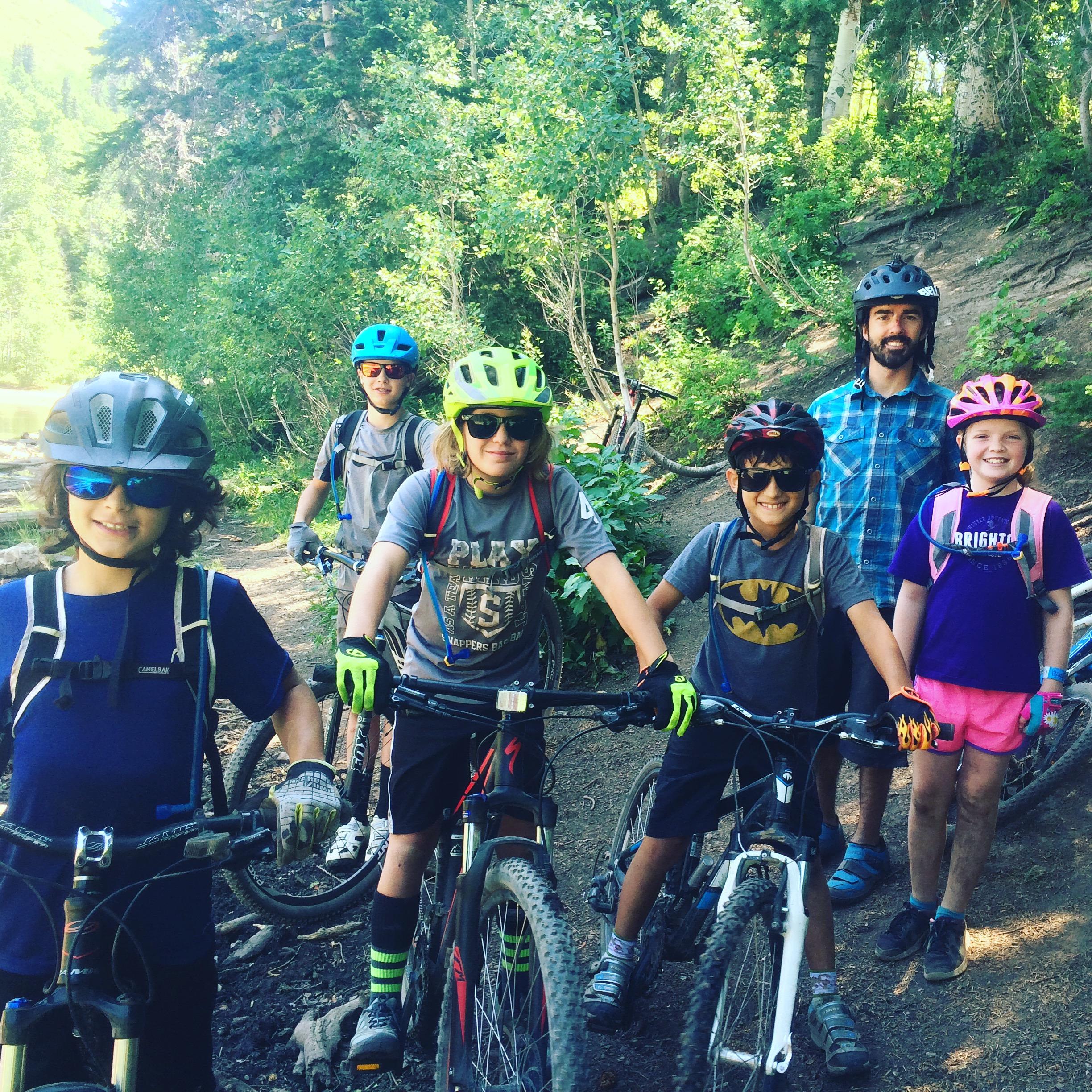 Giant Anthem X 29er 1: A group of six children and an adult, all wearing helmets and sunglasses, pose with their mountain bikes on a dirt trail surrounded by greenery. The children are smiling and dressed in casual biking attire, ready for an outdoor adventure.